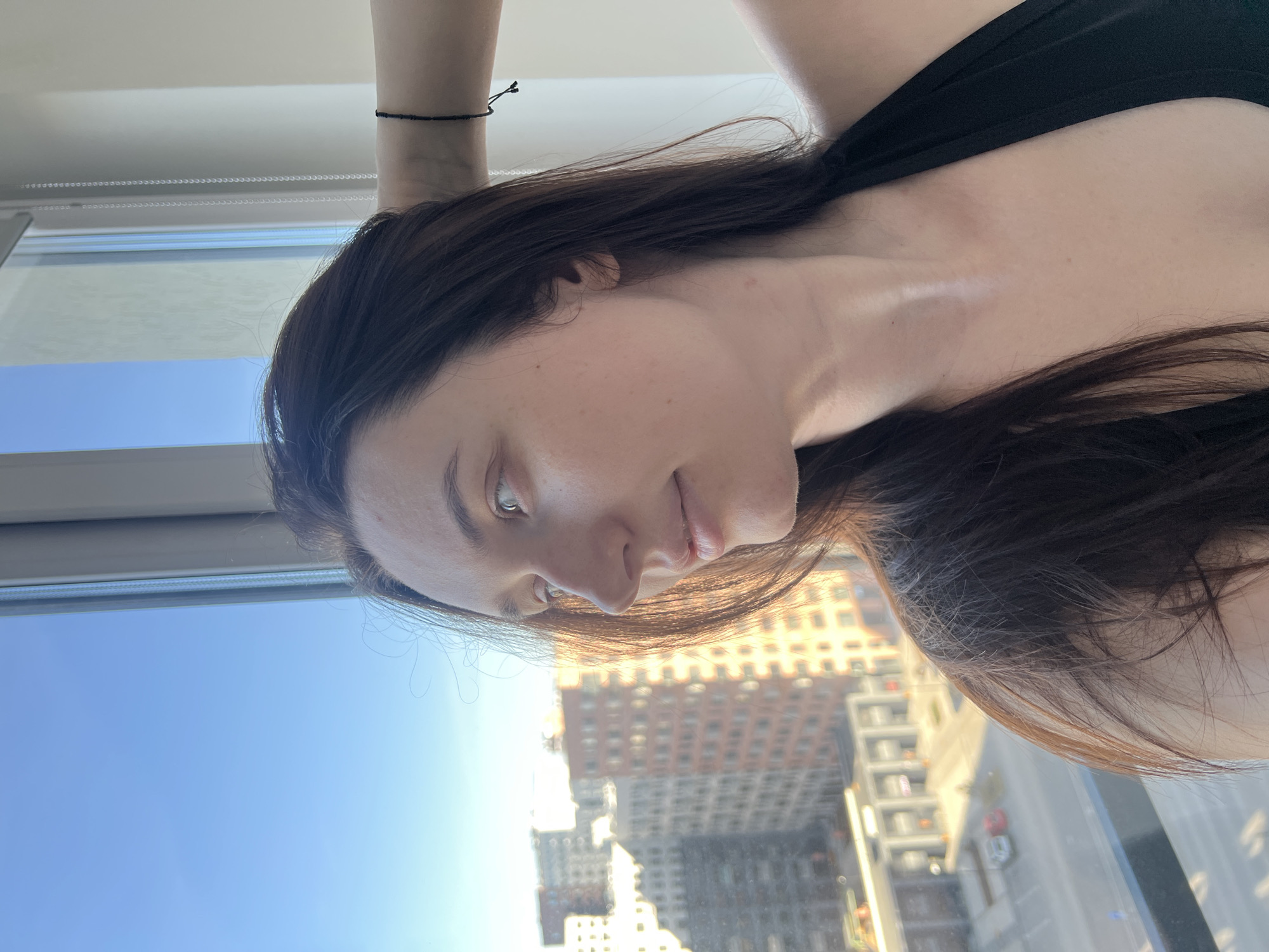 A young woman with long, dark brown hair and fair skin is looking out a window. She is wearing a black top, and a thin black bracelet is visible on her wrist. The window offers a view of a cityscape with tall buildings. A high-rise building with multiple windows is visible through the window. The blue sky indicates it is daytime. Based on the architecture and urban layout visible through the window, the picture was likely taken in Tyumen, Russia.