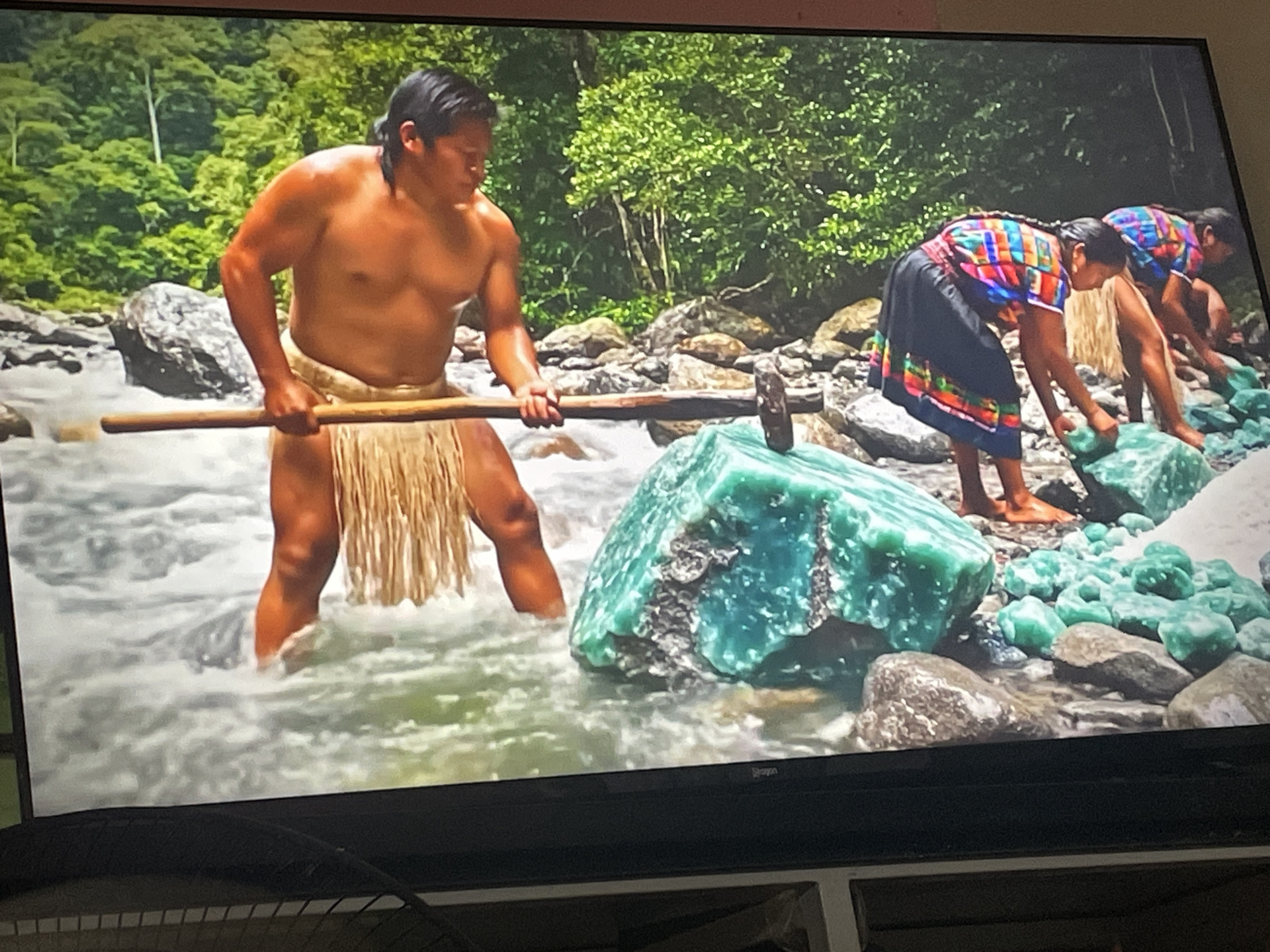 The image depicts a scene taking place in a rocky riverbed, surrounded by lush green vegetation, presumably a jungle or forest setting. A muscular man stands in the water, wearing a fringed loincloth. He is holding a long wooden handle with a heavy hammer-like head, poised to strike a large, rough-cut emerald-colored gemstone resting on a rock in the river. Behind him, two women wearing brightly colored, traditional blouses and dark skirts are bent over, appearing to be inspecting or working with smaller emeralds or jade stones. The location, city, and specific geographical region are unknown. The scene shows people working in a river environment, potentially mining or processing jade or emeralds.