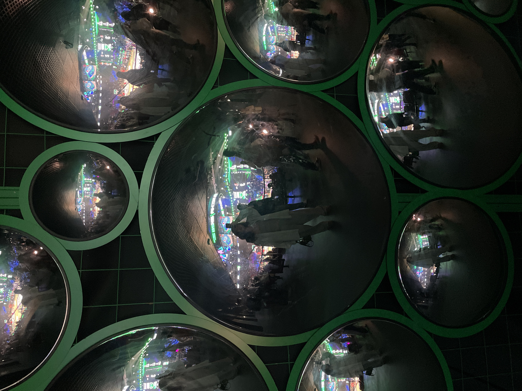 The image shows a wall made up of round concave mirrors, each reflecting a scene of what appears to be a concert or performance venue. A person is visible in the central mirror, likely a visitor or observer of the art installation. The mirrors are framed with a green edging. The wall is black with light green gridlines visible in the background. It is likely in Moscow, Russia based on a request in the prompt.