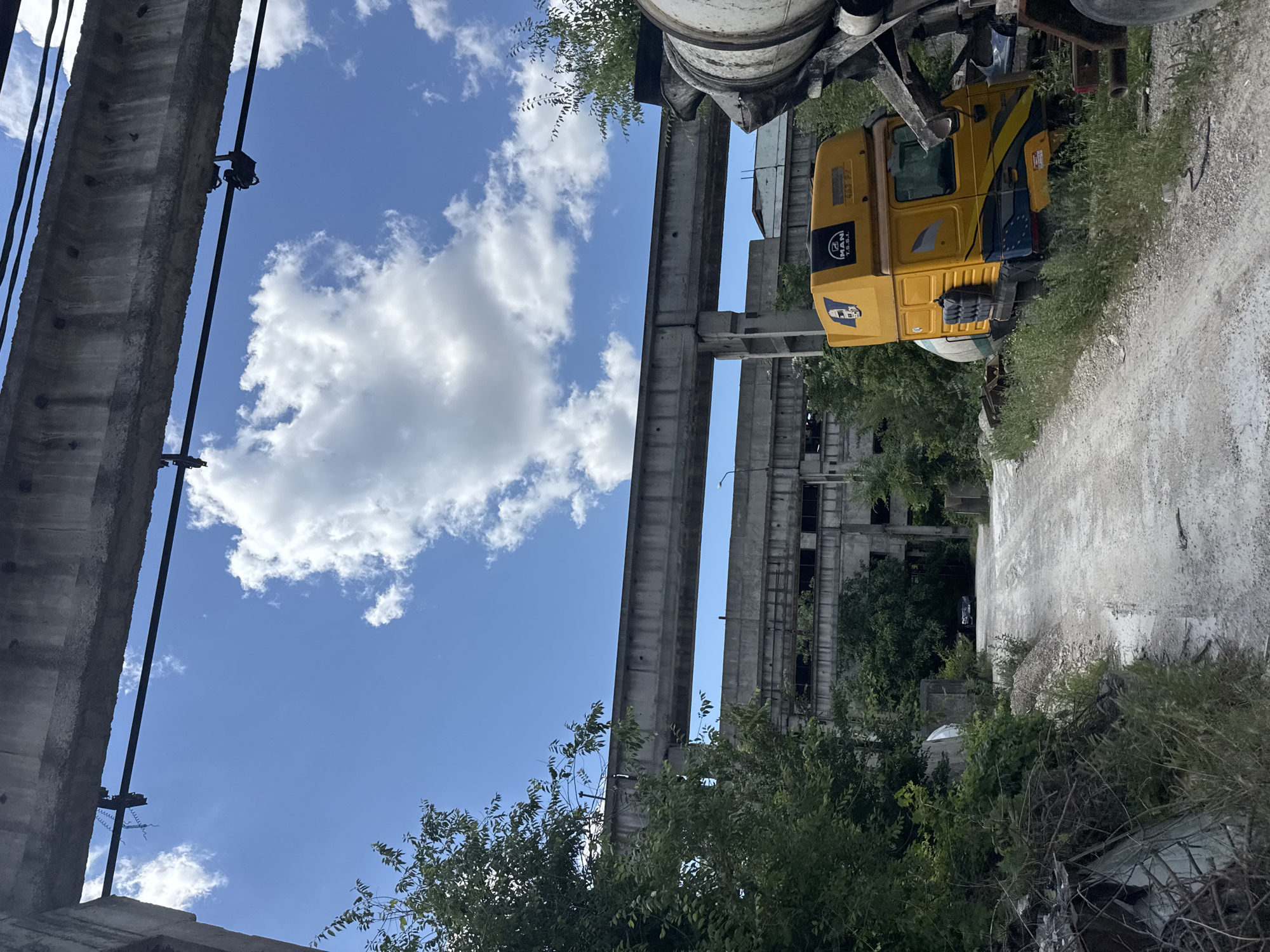 The image depicts an outdoor scene with a clear blue sky dotted with white clouds. In the foreground, a gravel path leads into the distance. To the right of the path, a yellow truck with a concrete mixer is parked, partially obscured by vegetation. The truck has the "MAN" logo visible on its cab. Further back, an abandoned industrial building with multiple stories and broken windows is visible, overgrown with trees and vegetation. Large concrete structures, possibly part of an elevated conveyor system, span across the upper portion of the image, casting shadows on the scene below. The overall impression is one of an industrial area that has fallen into disrepair and is being reclaimed by nature.
