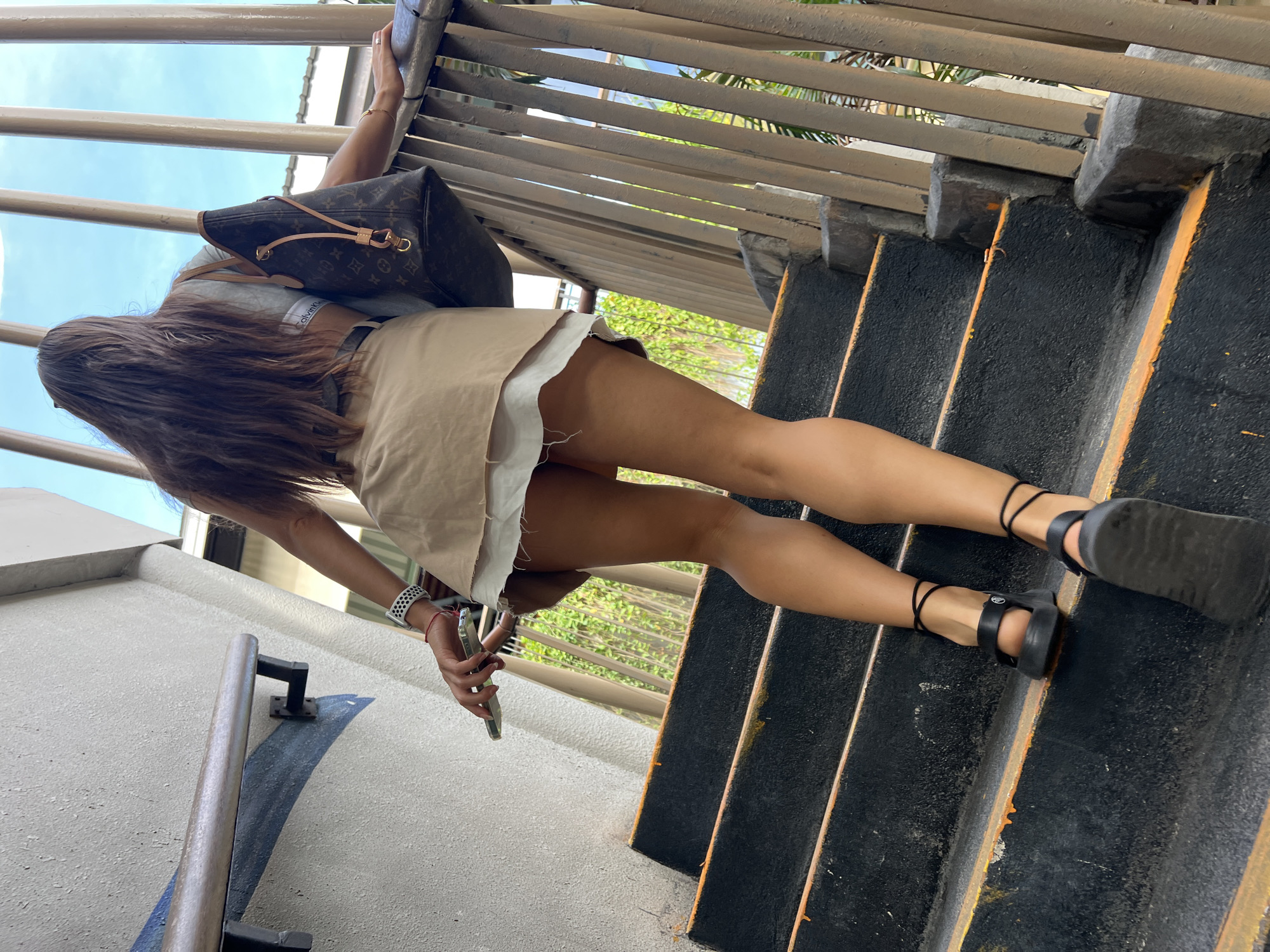 A woman is captured from behind, ascending a concrete staircase with black treads and yellow-orange risers. She has long, dark hair, is wearing a light-colored crop top, a tan skirt over white shorts, and black platform sandals with ankle straps. She is carrying a brown Louis Vuitton-patterned tote bag over her right shoulder and holding a silver smartphone in her left hand. The staircase is bordered by a metal railing on the right and a textured concrete wall on the left, which has a blue painted design. The background reveals a bright sky with vertical wooden or metal beams and lush green foliage. The image appears to be taken outdoors, likely in a public area.