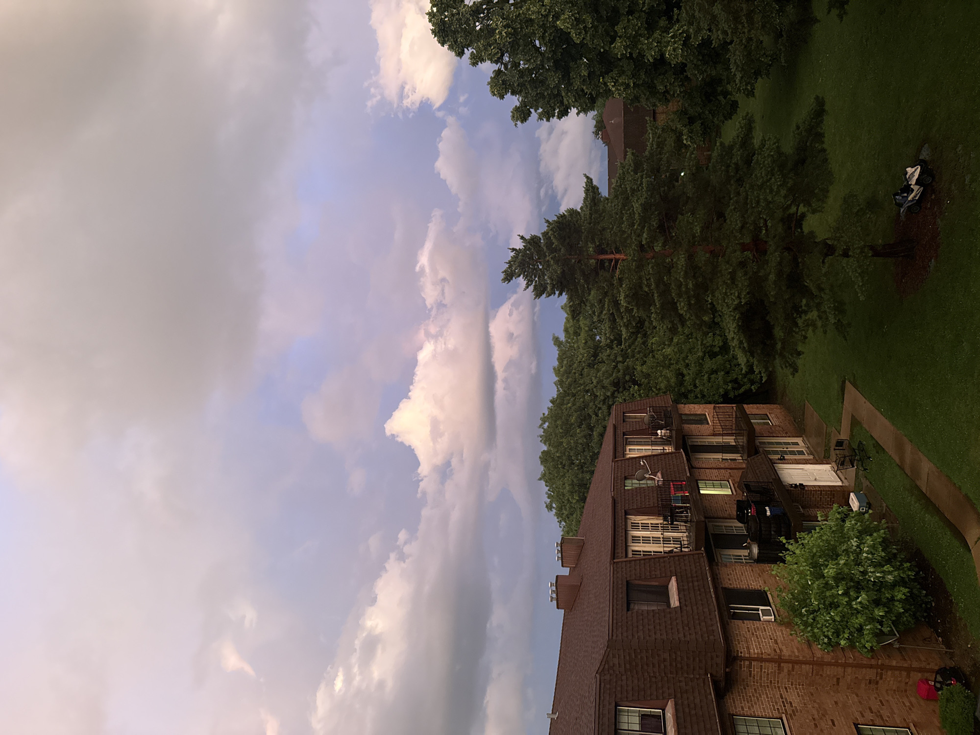 A high-angle, medium shot captures an apartment building with a brown, shingled roof and brick facade, situated amidst lush green trees under a dramatic sky filled with pink and purple-tinged clouds. The apartment building has multiple levels with visible windows, balconies, and chimneys. A tall pine tree stands prominently in the foreground to the right of the building, with a small utility vehicle parked at its base. The ground is covered in well-maintained grass, with a paved walkway leading towards the building. The scene appears to be in a residential area, possibly during twilight or a break in a storm, given the cloud formations and lighting.