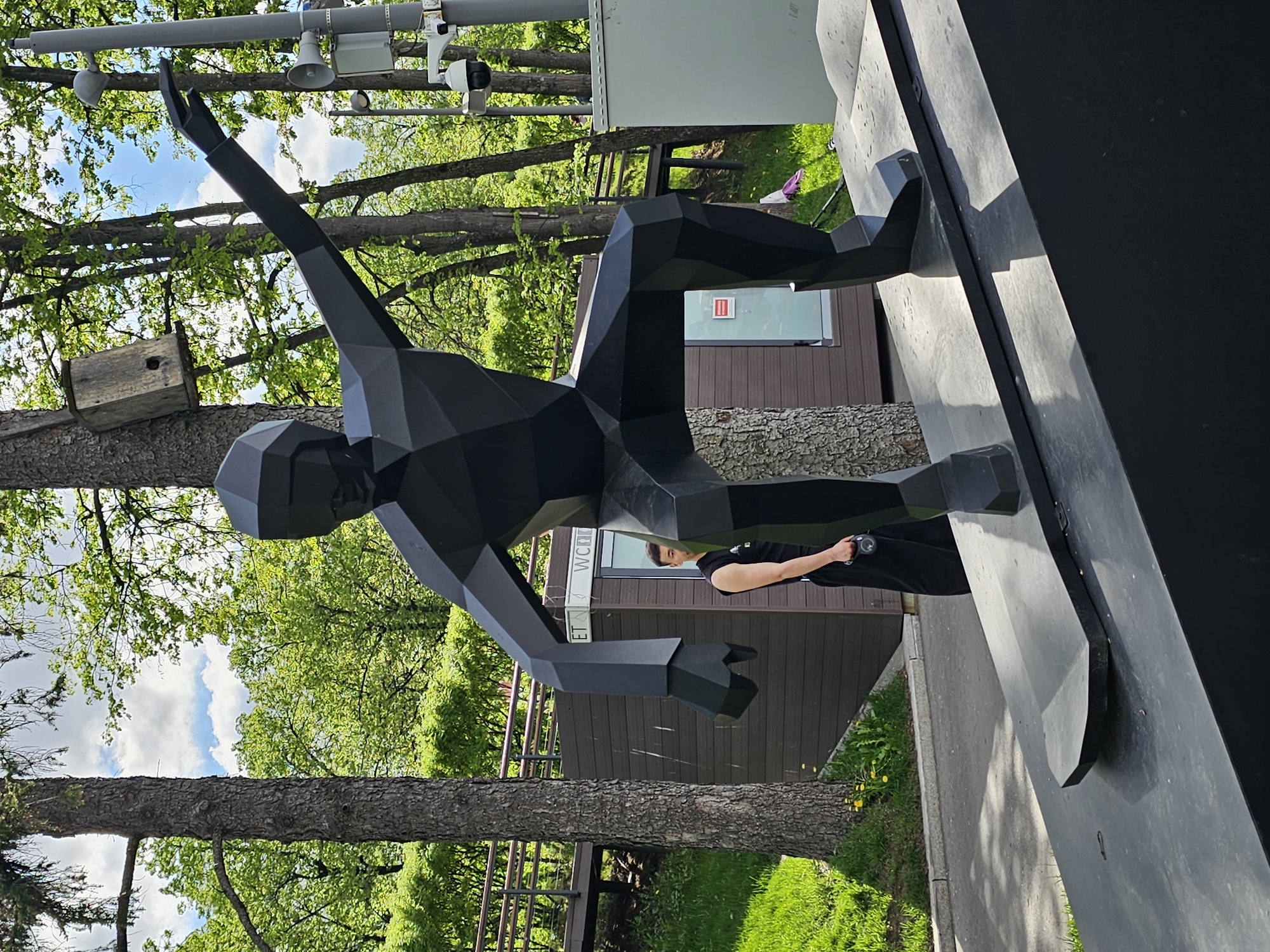 The image captures a sculpture of a stylized human figure with polygonal, angular features. The sculpture is predominantly black. One arm is raised toward a birdhouse in a tree. A person stands near the base of the sculpture. The location appears to be a park or green space in Moscow, Russia. The background features green trees, a partly cloudy sky, and structures reminiscent of public amenities or park buildings, including security cameras and a WC sign. The sculpture is positioned on a pedestal.