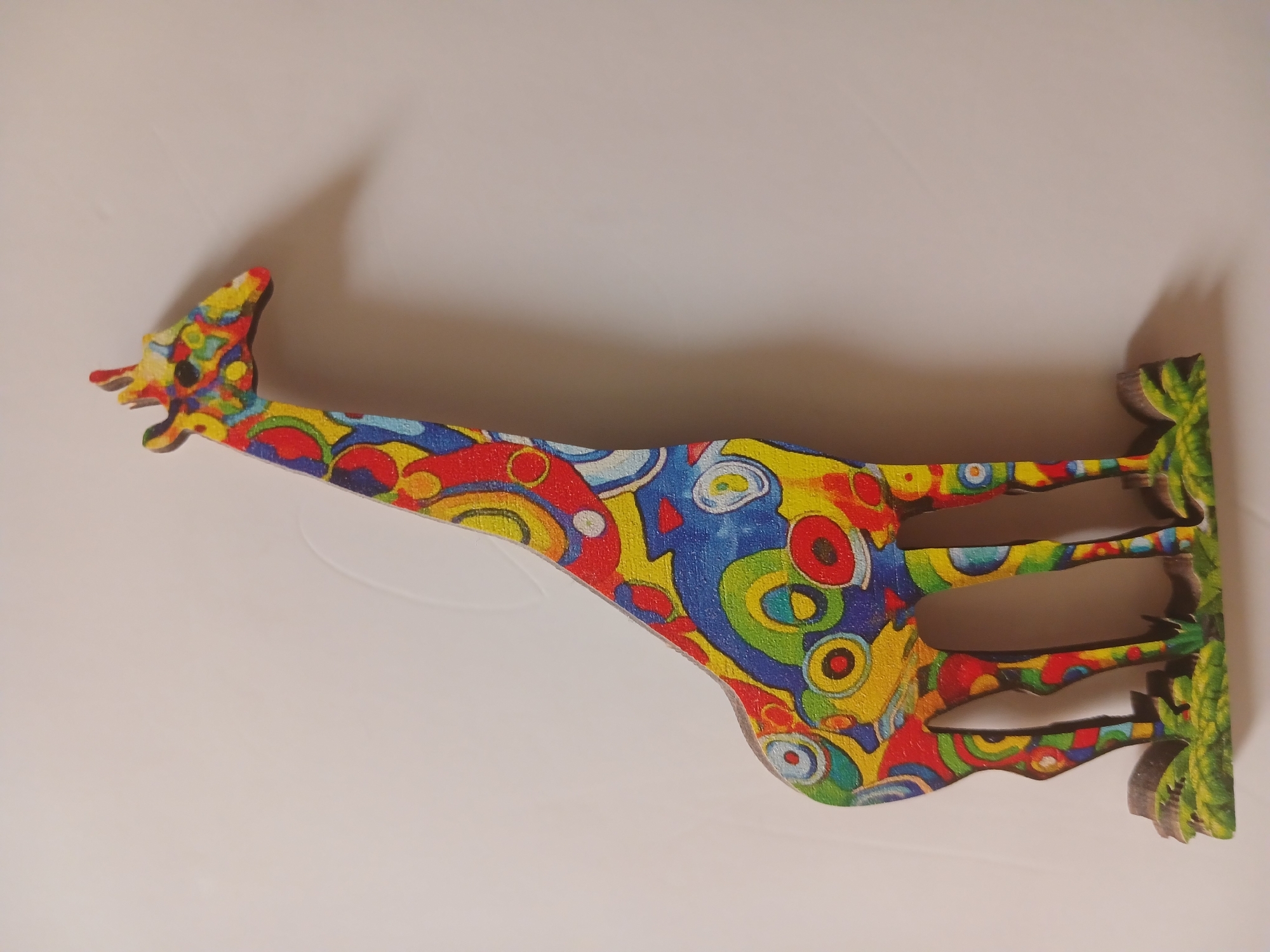 A vibrantly colored, stylized wooden giraffe figurine stands against a plain, off-white background. The giraffe is decorated with a psychedelic pattern of overlapping circles and swirls in red, yellow, blue, green, and white. It stands on a green base that resembles grass and rocks. The lighting casts a subtle shadow of the giraffe on the background to its right. The image does not provide any information about its location in the city of Sumy, Ukraine.