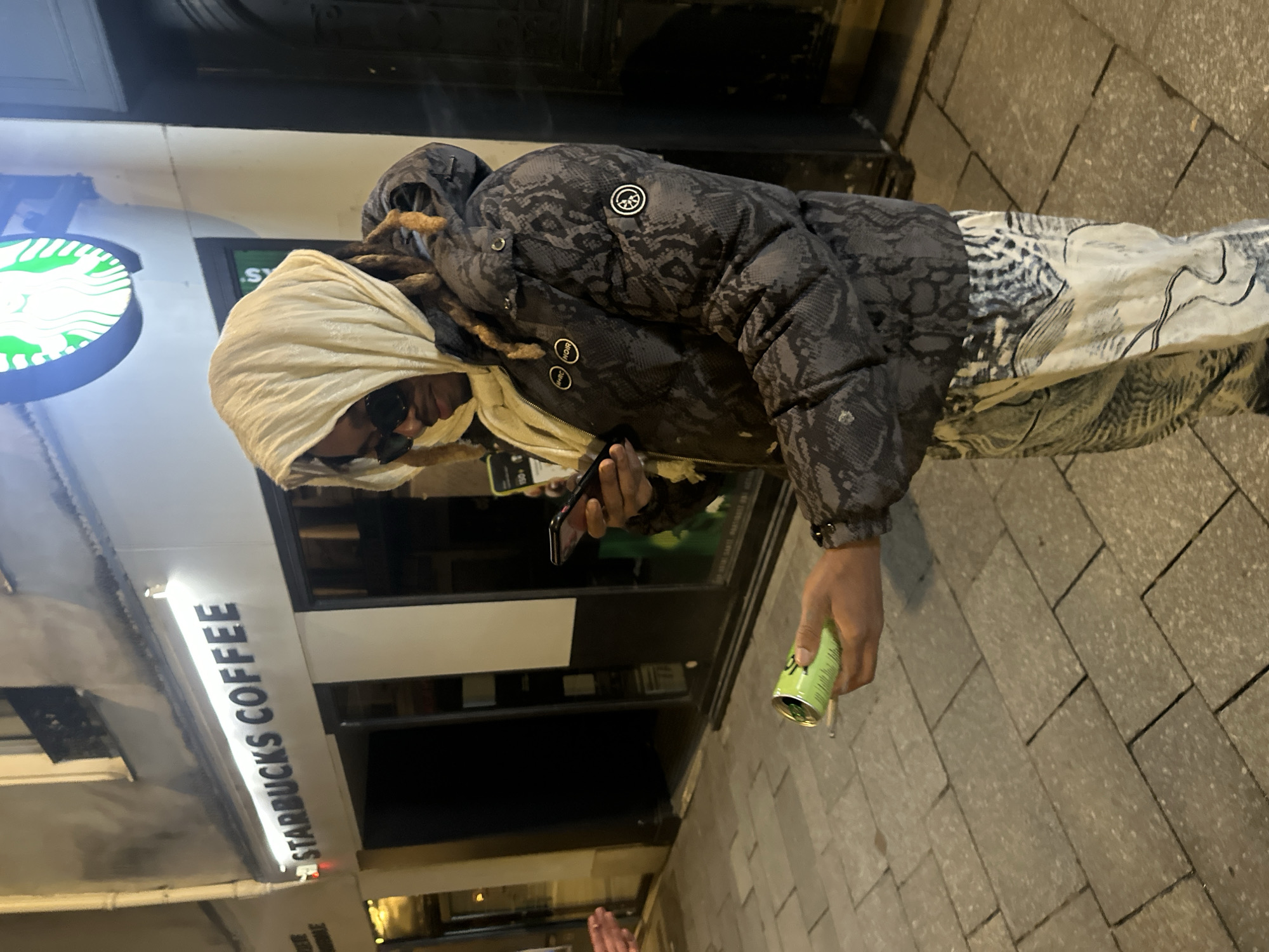 A person stands on a gray paved sidewalk outside a Starbucks Coffee establishment in Paris, France, at night. The individual is oriented slightly to the left, looking down at a smartphone held in their left hand. They wear a cream-colored fabric wrapped around their head, dark sunglasses, a dark gray puffer jacket with a subtle reptilian pattern (featuring "M+RC NOIR" and a peace sign-like circular patch), and light-colored pants with a dark, intricate pattern. In their right hand, they hold a light green aluminum can, partially obscured, with a rolled cigarette or joint resting on its rim. A human hand is visible extending into the frame from the far left. The background features the illuminated "STARBUCKS COFFEE" sign and a green circular Starbucks logo mounted on a white building facade with dark-framed windows and an entrance. A vertical drainpipe and an upper-story balcony are also visible on the building.