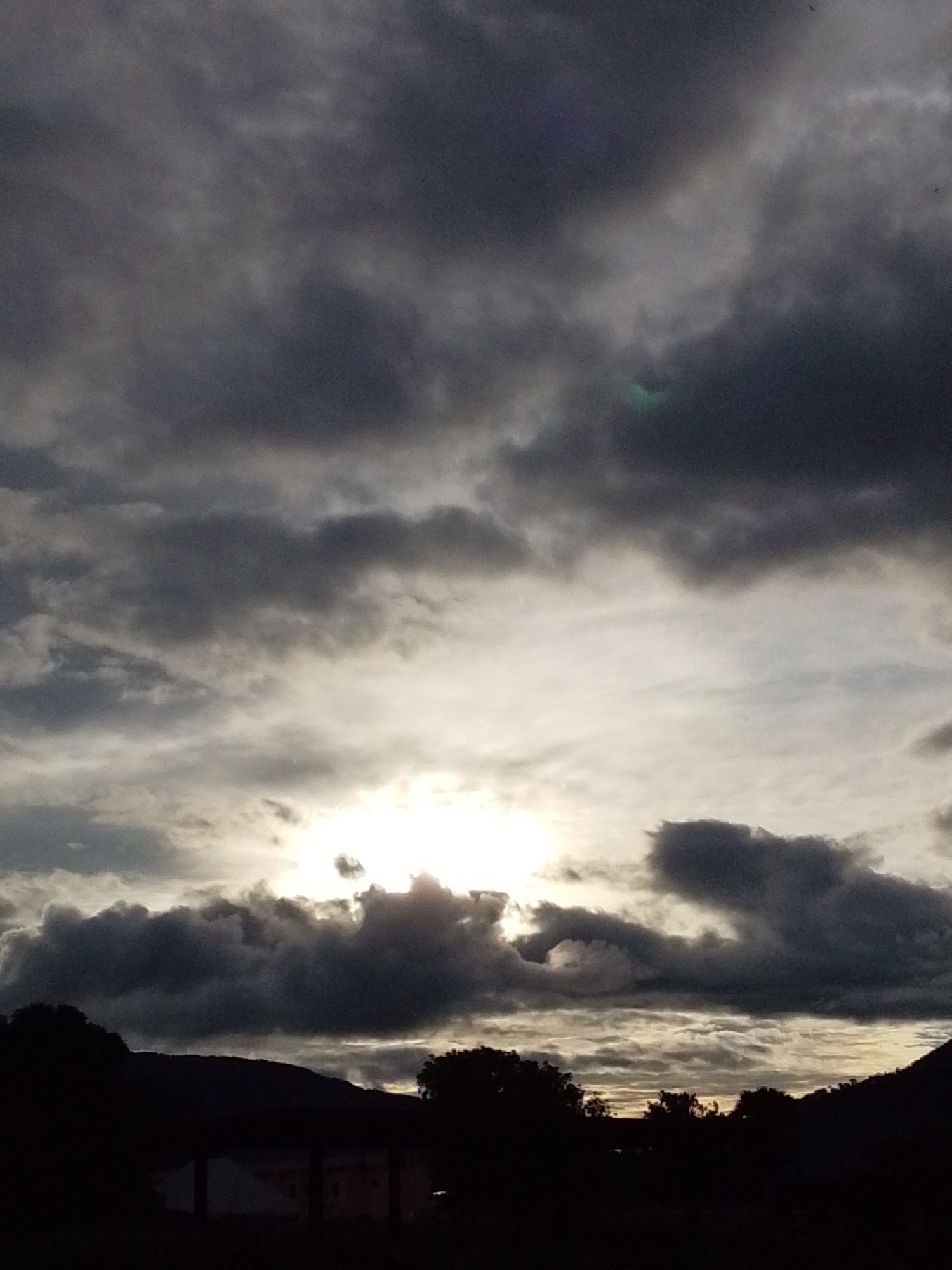 The image depicts a dramatic sky over a silhouetted landscape, likely taken at dusk or dawn. Dark, heavy clouds dominate the upper portion of the frame, with shafts of bright, diffused sunlight breaking through in the middle. The sun itself is largely obscured by cloud cover, creating a glowing, hazy effect. Below the sky, a dark, indistinct line of hills or mountains forms the horizon. In the foreground, the silhouettes of trees and what appear to be rooftops of buildings are visible against the bright sky. The overall impression is one of a moody, atmospheric natural scene. This scene is consistent with the natural environment of Serti, Nigeria, characterized by its varied topography and seasonal weather patterns which can produce such skies.