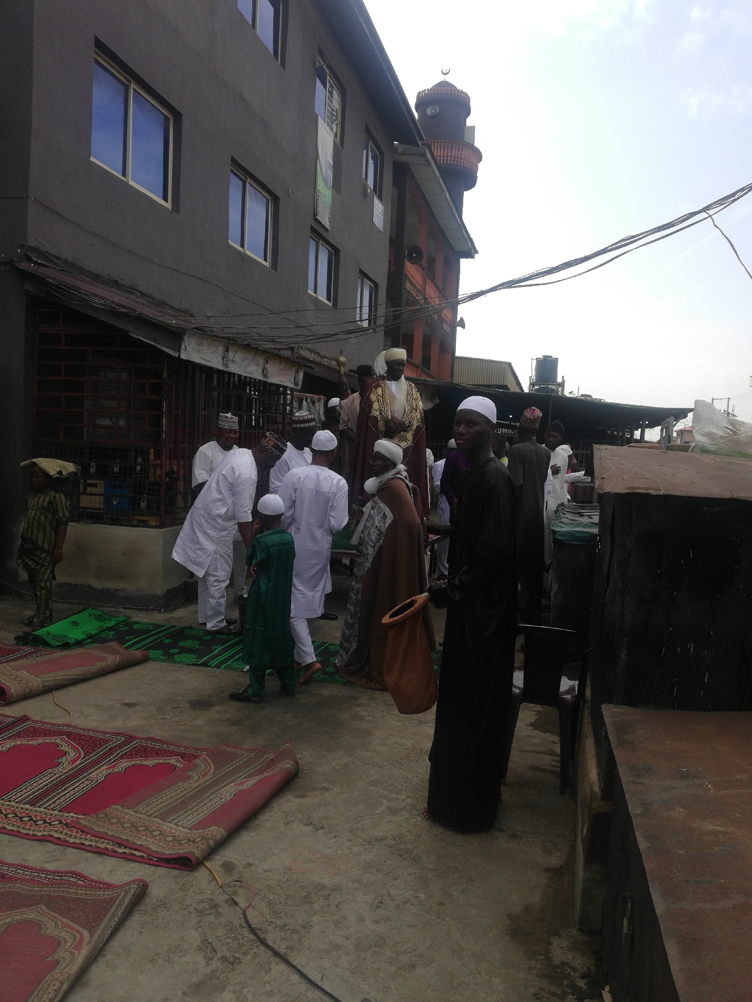 The image depicts a gathering of people outside a building that appears to be a mosque in an unidentified urban area. Several individuals are dressed in traditional Muslim clothing, including white robes and caps. One person is wearing a more ornate garment, suggesting a position of leadership or status. Children are also present, dressed in green outfits. Prayer rugs are laid out on the ground, indicating preparations for prayer. The mosque's minaret is visible in the background. Overhead, there are numerous wires spanning the area. The environment includes a mix of residential and commercial elements, as indicated by the building's construction and the presence of what appears to be a storefront.