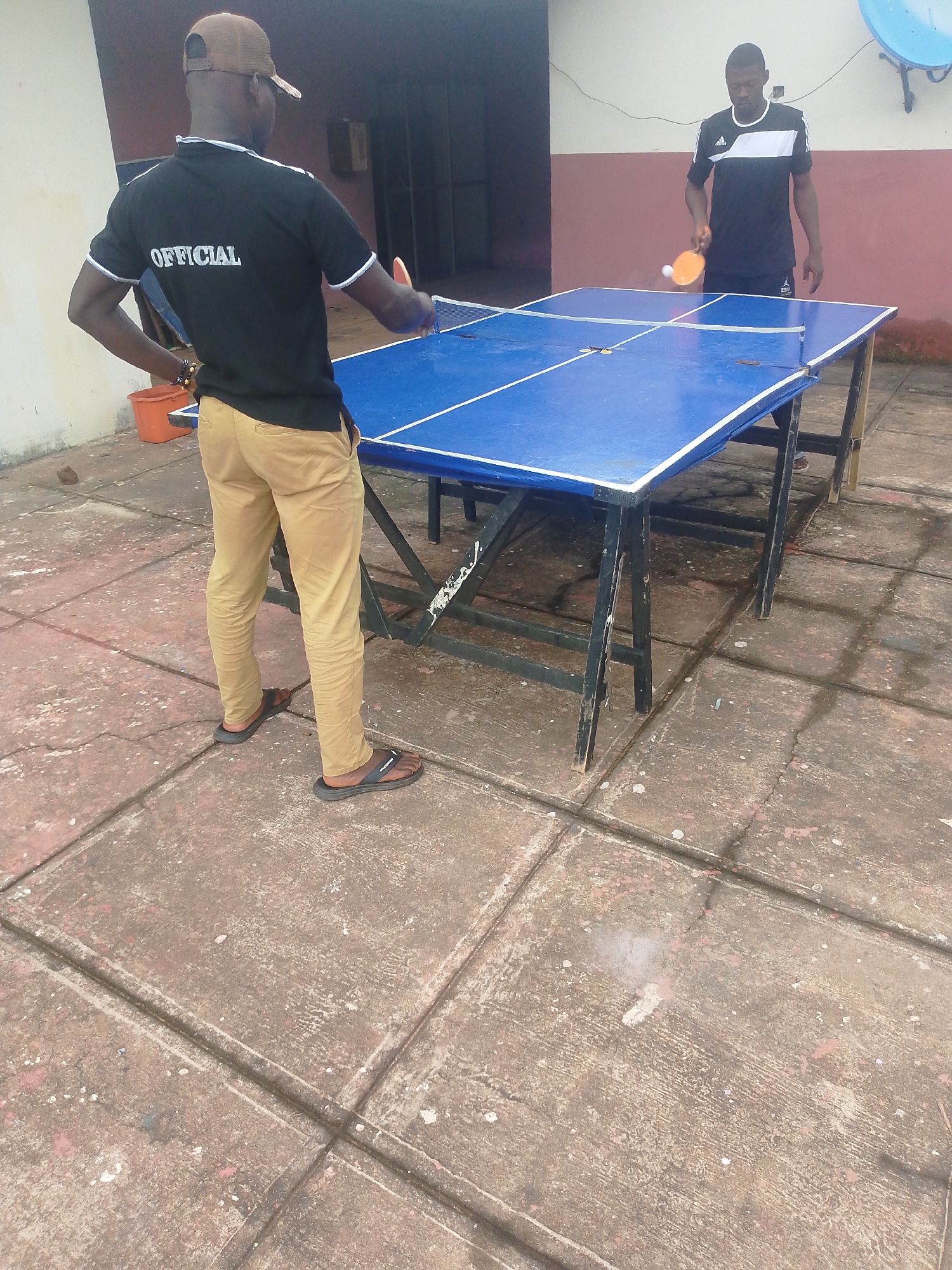 Two men are playing table tennis outdoors on a concrete surface in Agbani, Nigeria. The table is blue with white lines and a net. The man on the left wears a black shirt with "OFFICIAL" written on the back, tan pants, and a brown cap. He is holding a ping pong paddle. The man on the right wears a black and white athletic shirt and dark pants. He is also holding a ping pong paddle and is in the process of hitting a ping pong ball. A large, blue satellite dish is visible on the wall behind them. The environment appears to be a courtyard or a paved outdoor area.