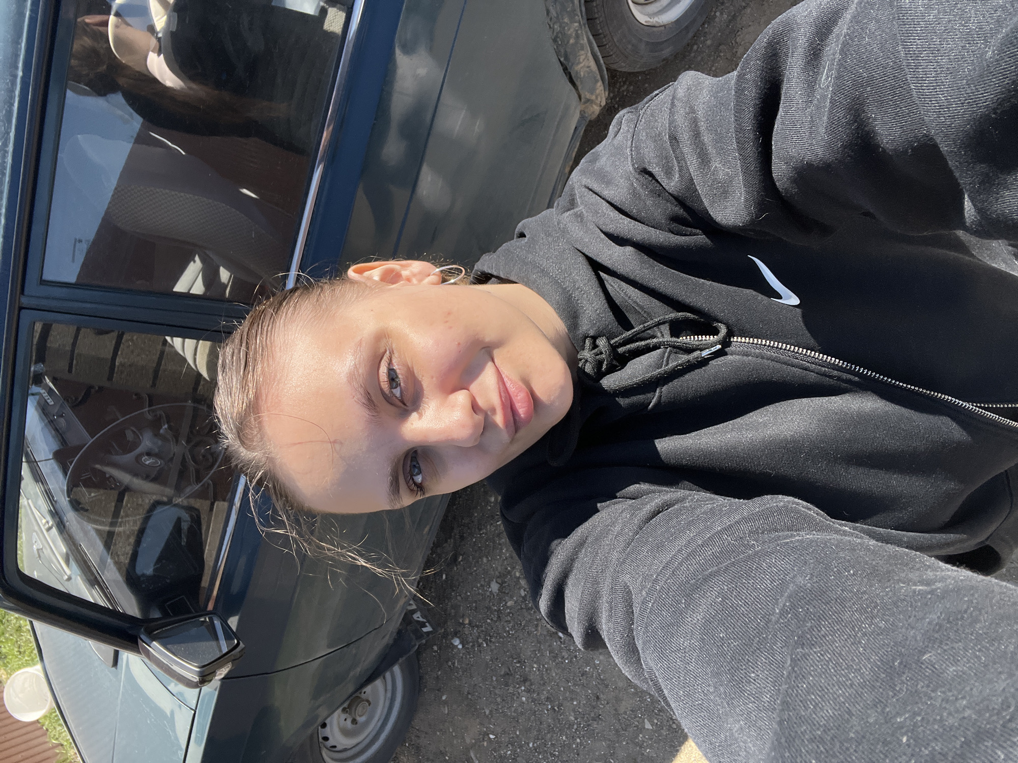 A selfie taken by a person with fair skin, light brown hair pulled back, and blue eyes. The person is wearing a black Nike zip-up hoodie. The person is standing next to a dark green car. The car has a square side mirror and white-rimmed tires. The setting appears to be outdoors on a gravel or dirt surface. It is daytime. The car's steering wheel is visible through the driver's side window. There are no visible details confirming the location as Ольховка, Russia.