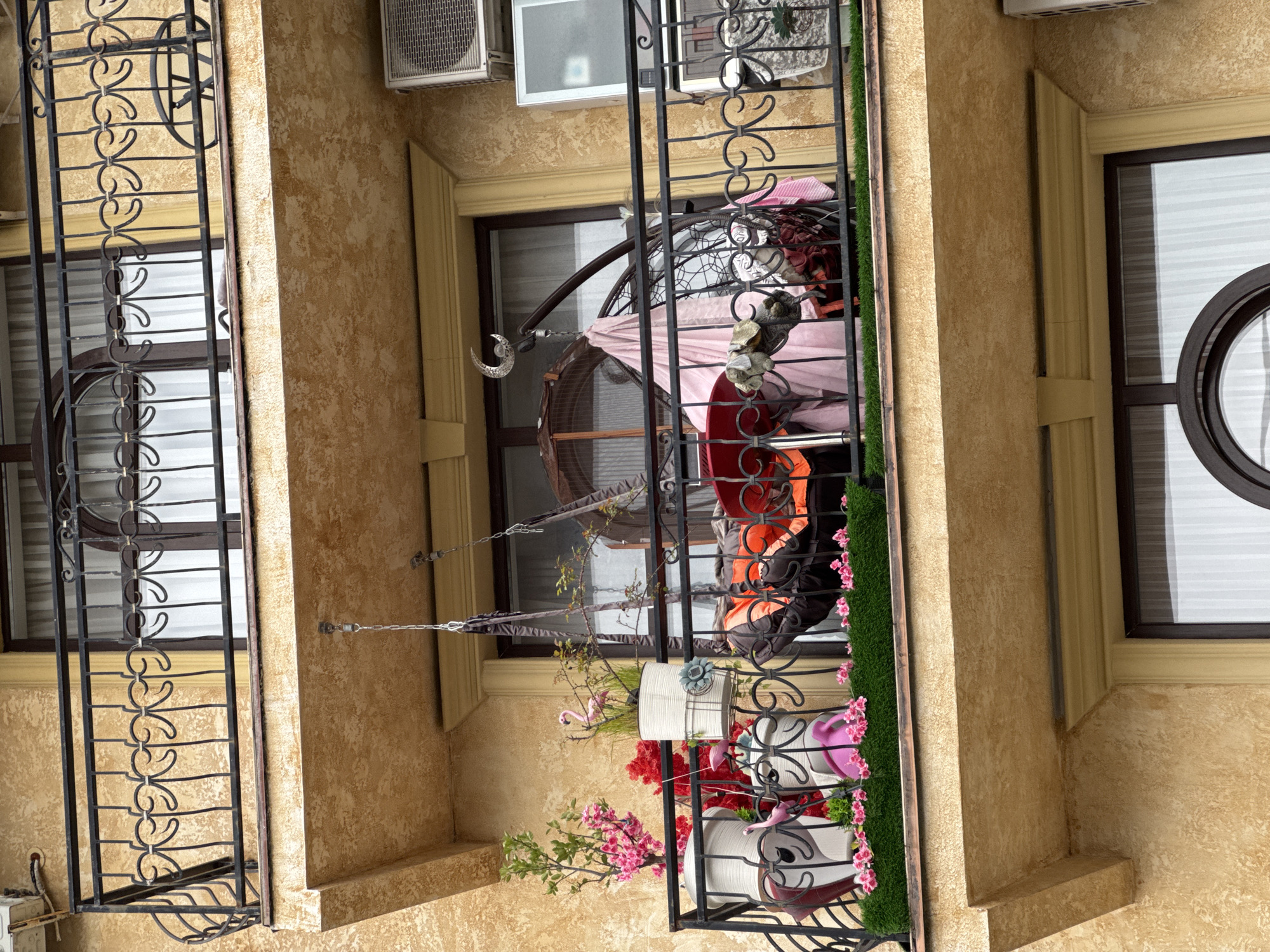 This image captures a close-up, upward-angled view of a building facade, likely an apartment complex or hotel, in Sirius, Russia. The building is constructed with a textured, light brownish-yellow stucco finish.
The main focus is on two balconies and several windows. On the left, a narrow balcony features an ornate black wrought-iron railing with scrollwork patterns. Behind it, a dark-framed window is partially covered by white horizontal blinds, and a circular decorative element is visible within its lower pane.
The central and largest balcony also has a black ornate railing, topped with a strip of artificial green turf and adorned with artificial pink flowers. Inside, a circular, dark-framed structure, possibly a hanging chair or swing, is visible, draped with pink fabric. Red and orange fabric items, likely cushions or clothing, are also present. A silver crescent moon ornament hangs on chains from the wall, leading towards the swing. Several white plant pots are arranged along the railing, containing more artificial pink flowers and small pink flamingo figurines. Above this balcony, an outdoor air conditioning unit is mounted on the building wall.
To the right, a similar dark-framed window with a circular detail is visible, also with internal blinds. The overall scene suggests a decorative and personalized outdoor living space. The lighting indicates it is daytime. No people are visible in the image, and there is no visible text.