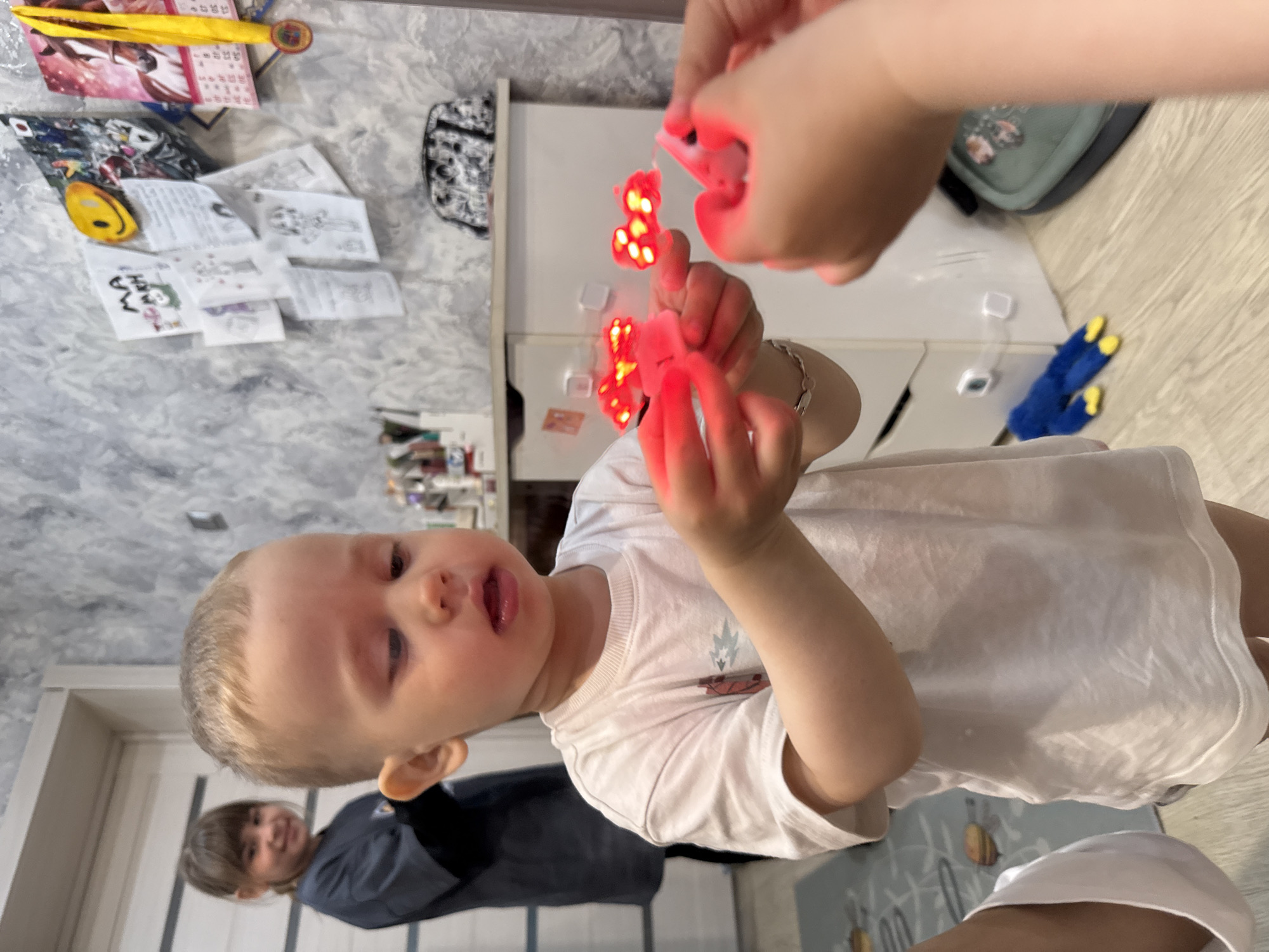 A young child in a white t-shirt is holding two red, heart-shaped light-up toys. Another person's hand is visible, also holding a similar toy. A girl stands in the background wearing a dark shirt. The location appears to be indoors, possibly a residence, with a wall decorated with children's drawings and a calendar. A white cabinet and flooring are visible. While Rusanovo, Russia is specified as the city, the image provides no specific details to confirm this location.