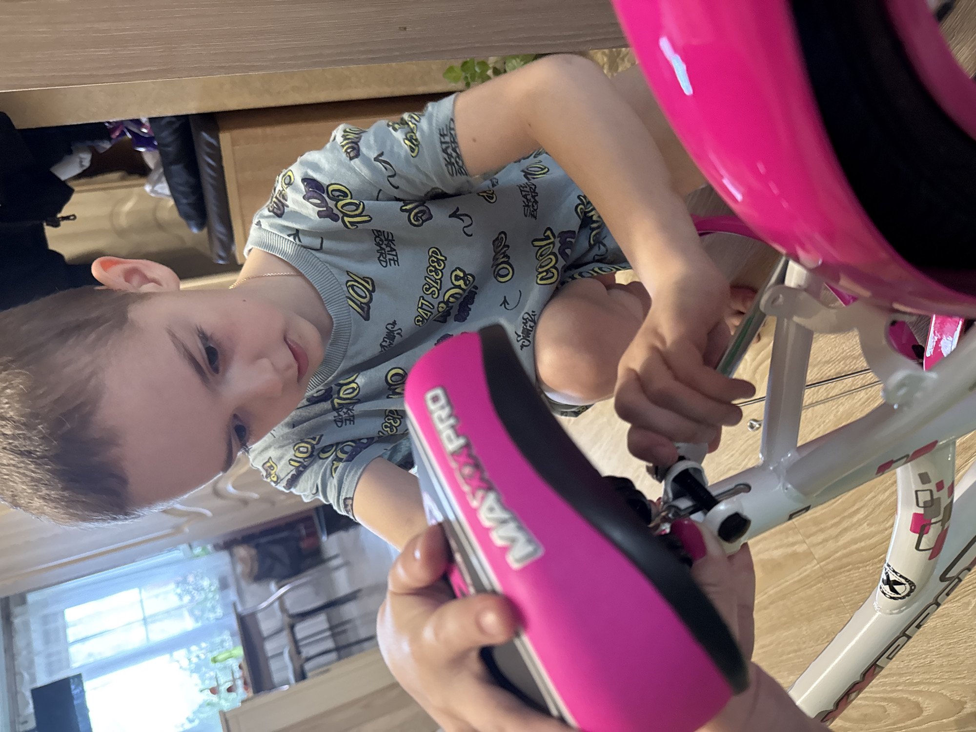 A child is shown indoors assembling a pink and white bicycle. The child wears a light blue printed t-shirt. The bicycle seat, labeled "MAX PRO," is pink and black. The bicycle has a pink helmet on top. The interior space is illuminated by natural light from a window, and the floor is wooden. The location mentioned is unrelated to the image content.