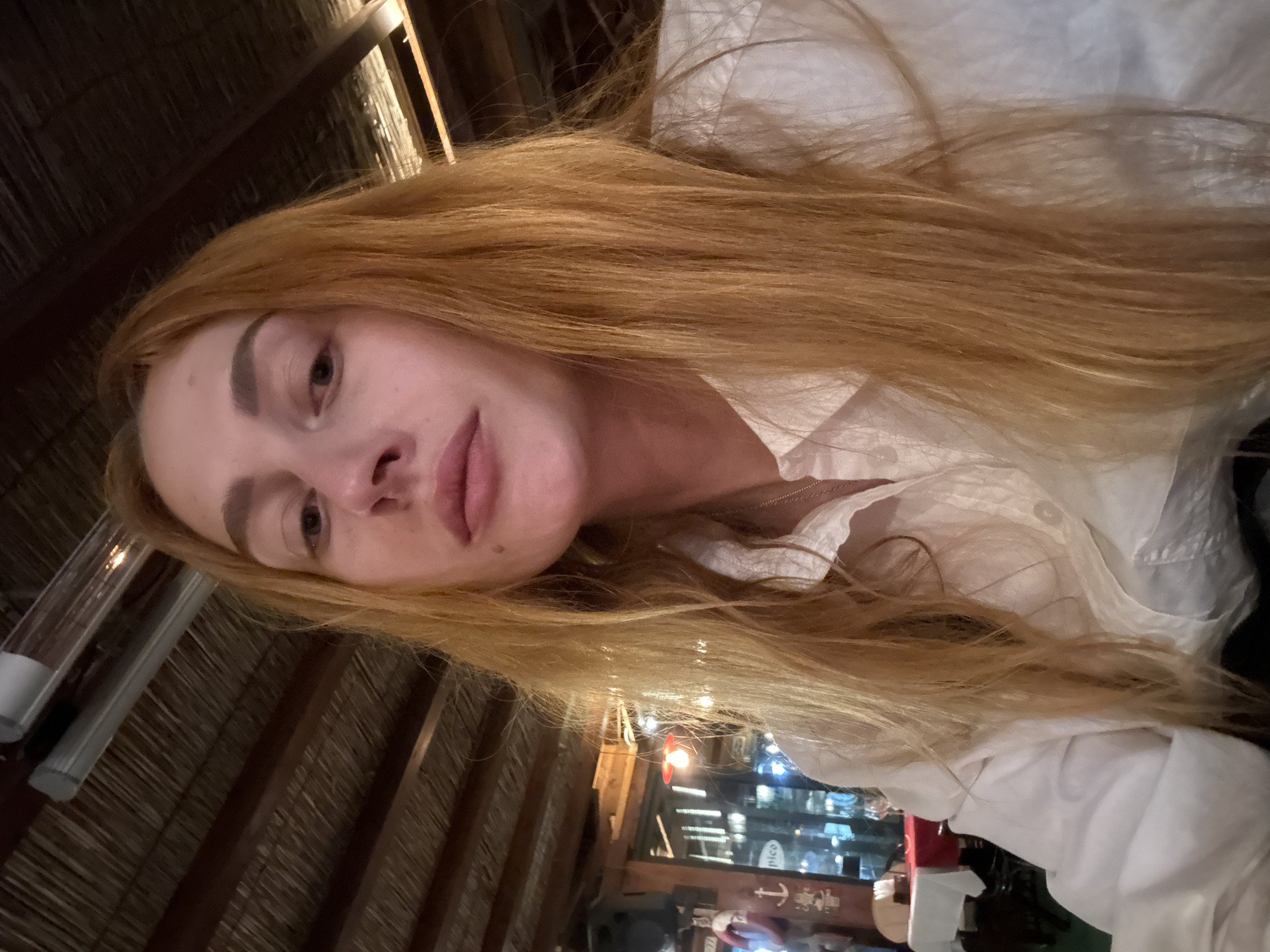 A person with long red hair is the primary subject of the photo. They are wearing a white collared shirt. The background reveals that the photo was taken indoors, likely in a restaurant or bar setting, in Budva, Montenegro. The building’s ceiling shows exposed beams. Lights and reflections are visible through the glass windows behind the person, displaying other structures, including a building with an anchor symbol in its window. The overall lighting suggests that the photo was taken in the evening.