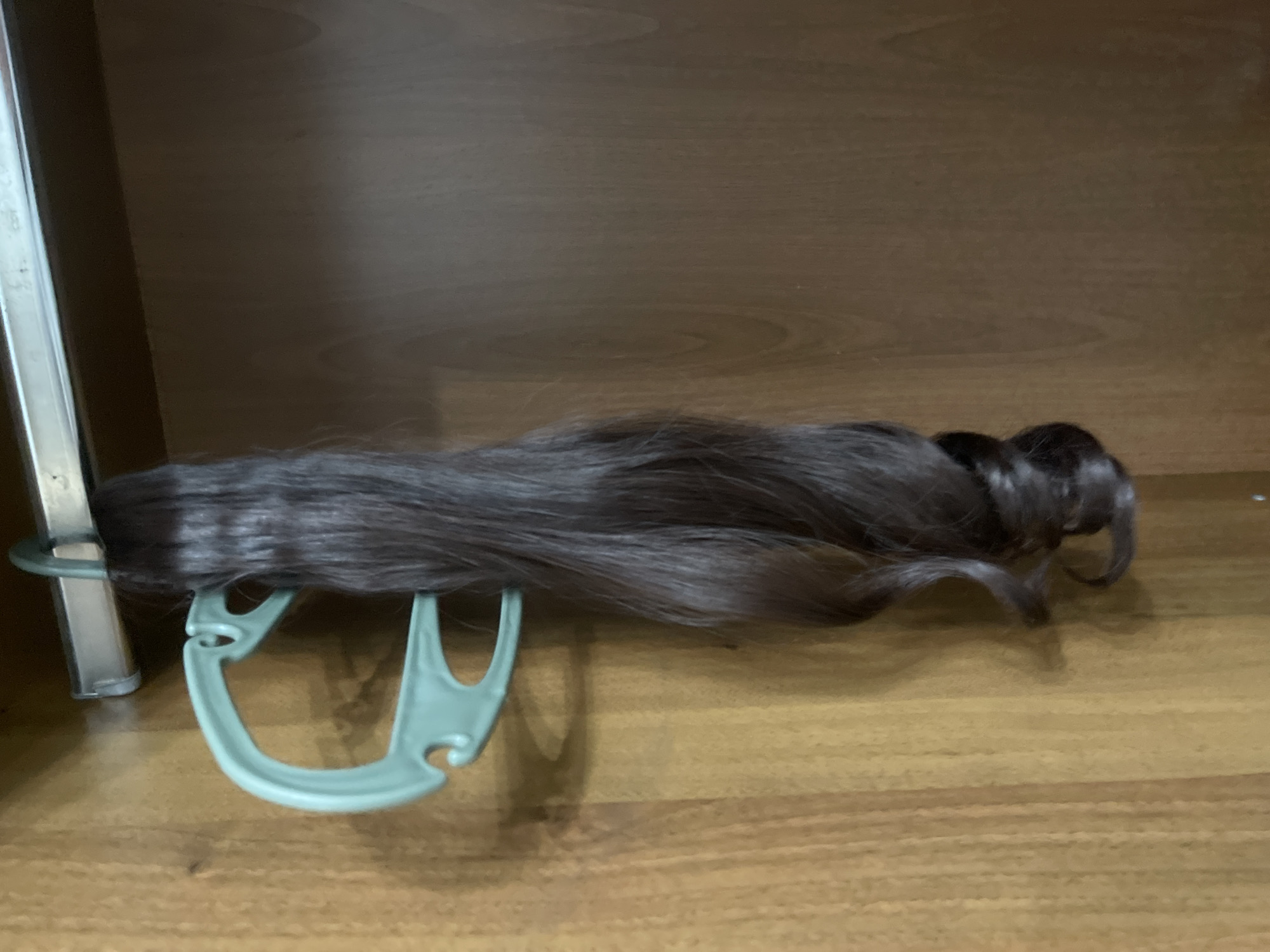 A dark brown wig is draped over a pale green plastic wig stand, which is resting on a wooden surface, likely a shelf. The wig is long and appears to be wavy, with a slight curl at the ends. The wig stand has a U-shaped design with an additional hook on one side. To the left of the wig stand, a portion of a silver metal object, possibly part of a closet or rack, is visible. The background is a plain, wooden surface that appears to be part of a shelf or cabinet. The lighting suggests an indoor setting, possibly during the day. There is no visible text in the image.