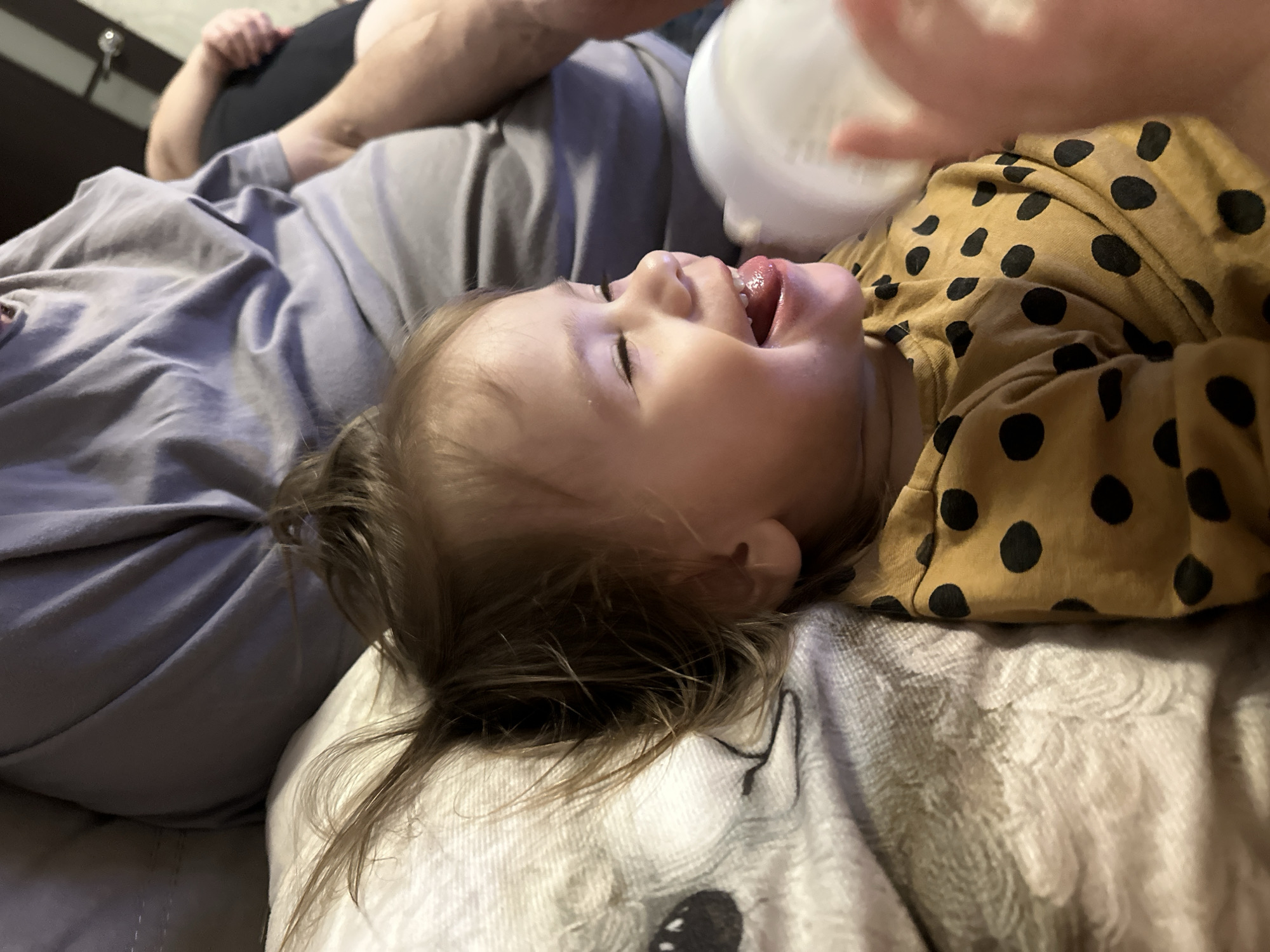 This intimate indoor photograph, set in Uva, Russia, captures a close-up moment of a young child being fed. The child, appearing to be a toddler with light brown hair, is lying on their back on a light-colored, patterned blanket or pillow. They are dressed in a cheerful mustard yellow top with prominent black polka dots. Their facial expression is one of pure joy and anticipation: eyes closed in a moment of contentment, mouth wide open, and tongue playfully extended, reaching towards a white feeding bottle held just above their face by an unseen hand.
Partially visible in the background is the upper body of an adult, wearing a grey t-shirt over what appears to be a black layer, suggesting the child is resting comfortably alongside them, possibly on a bed or sofa. The soft, ambient lighting points to an indoor setting, likely a home, and creates a peaceful and tender atmosphere around the feeding activity. The child's wide smile and eager posture convey happiness and comfort. No discernible text is present in the image.