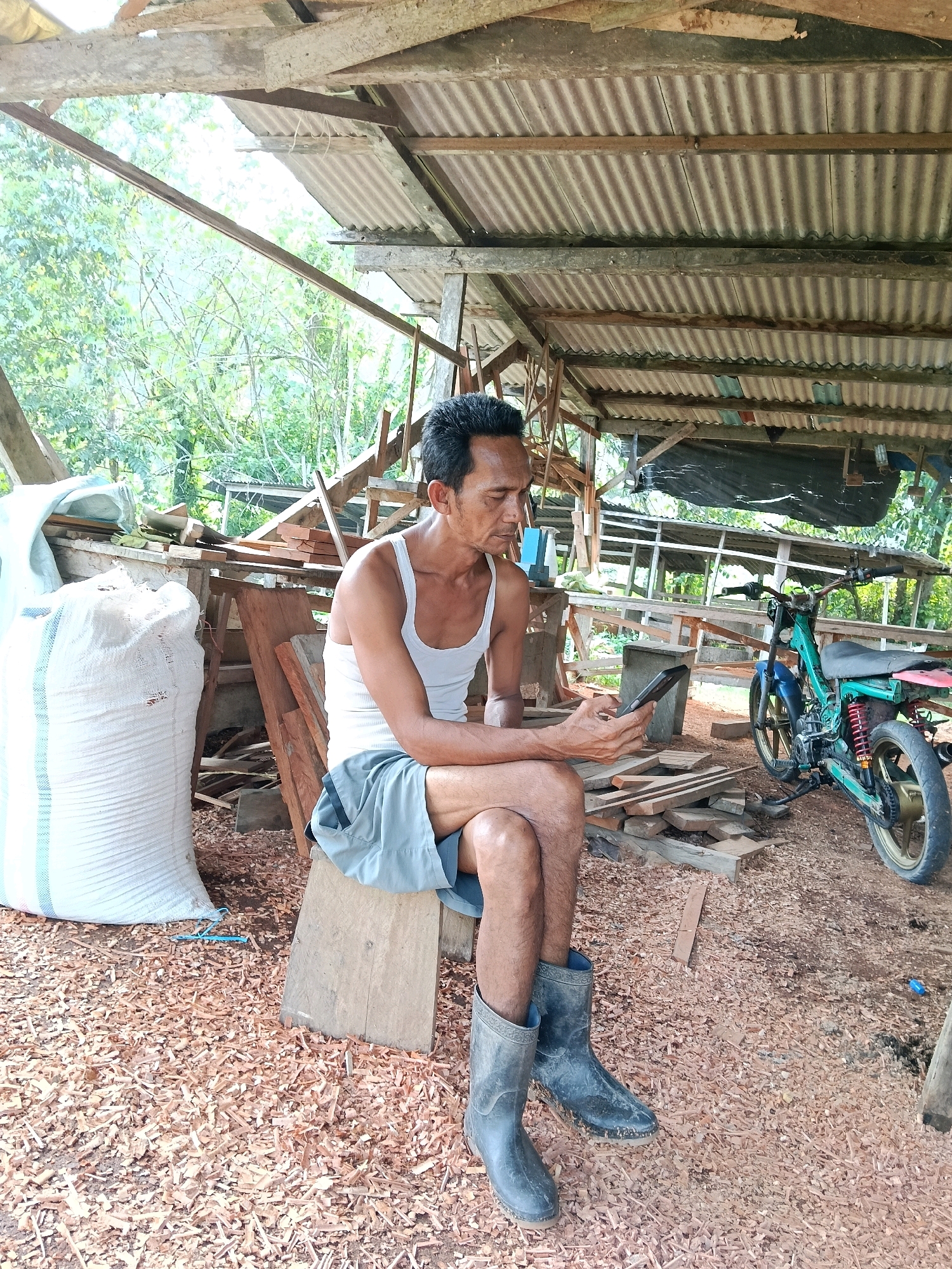 An Indonesian man is sitting on a wooden stool in a workshop, looking at his smartphone. He is wearing a white tank top, grey shorts, and black rubber boots. The workshop is filled with woodworking debris, including piles of wood shavings on the ground and stacks of lumber. A large white sack is propped up to his left, and a green motorcycle is parked behind him. The workshop appears to be open-air, with a corrugated metal roof supported by wooden beams. Trees and foliage are visible in the background, suggesting an outdoor or semi-outdoor setting. The scene is located in Muara Siberut, Indonesia, a town known for its woodworking and traditional crafts.