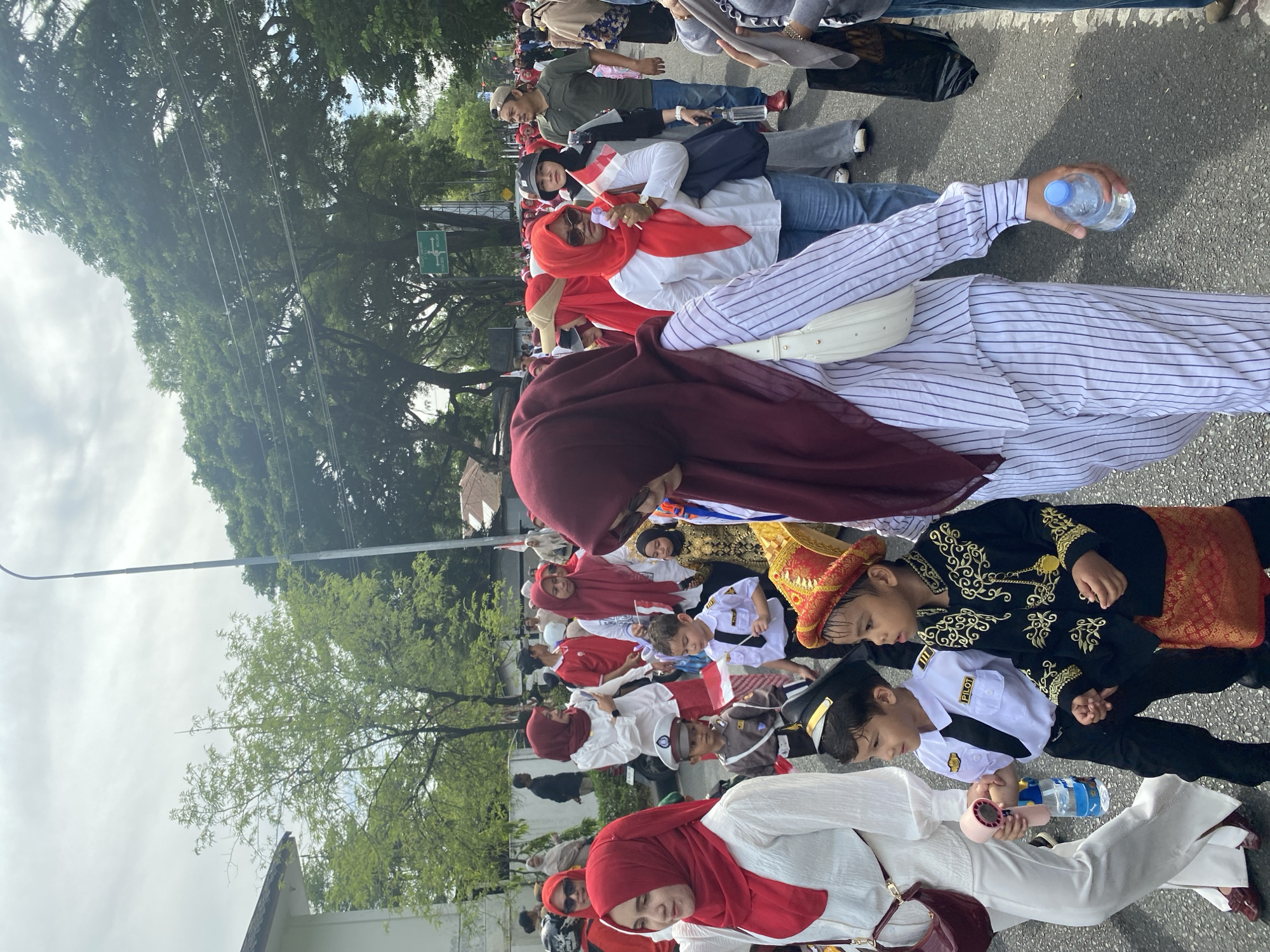 The image captures a street scene in Banda Aceh, Indonesia, where a parade is in progress. A group of people, including adults and children, are walking on a paved street. Many are wearing red and white attire, with some adults wearing hijabs. Several children are dressed in costumes, including one in a pilot uniform and another in a traditional ceremonial outfit. Some individuals are holding Indonesian flags. The environment includes trees lining the street, a building on the left, and a lamppost. The sky is overcast. The overall mood appears festive, likely in celebration of a national holiday.
