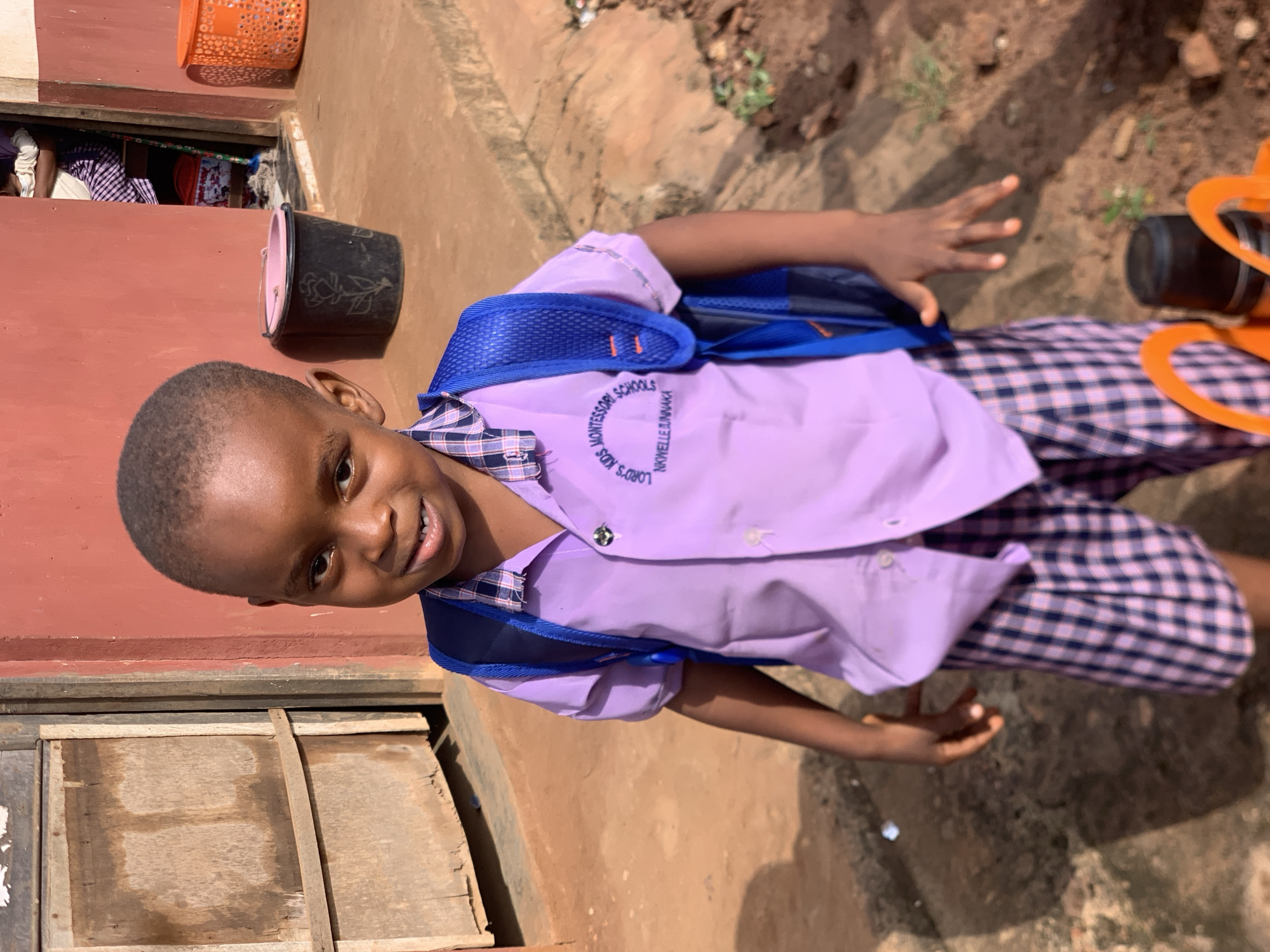 A young Black boy stands outdoors, facing forward, wearing a light purple collared shirt with a checkered collar and cuffs, and matching checkered shorts. He has a blue backpack on. The shirt has an embroidered circular logo on the left chest area that reads "LORD'S KIDS MONTESSORI SCHOOLS" with "NKWELLE-UNNÁKA" below it. The boy is positioned in front of a reddish-brown textured wall, with a wooden door visible to his left. The ground is dirt and appears uneven. In the background, there are several objects including a black bucket with a pink lid and a rose design, and an orange plastic basket. There is also an open doorway showing a person in a checkered skirt and a white top. The scene is likely in Ukwu-Abwa, Nigeria, given the environment and common school uniform styles.