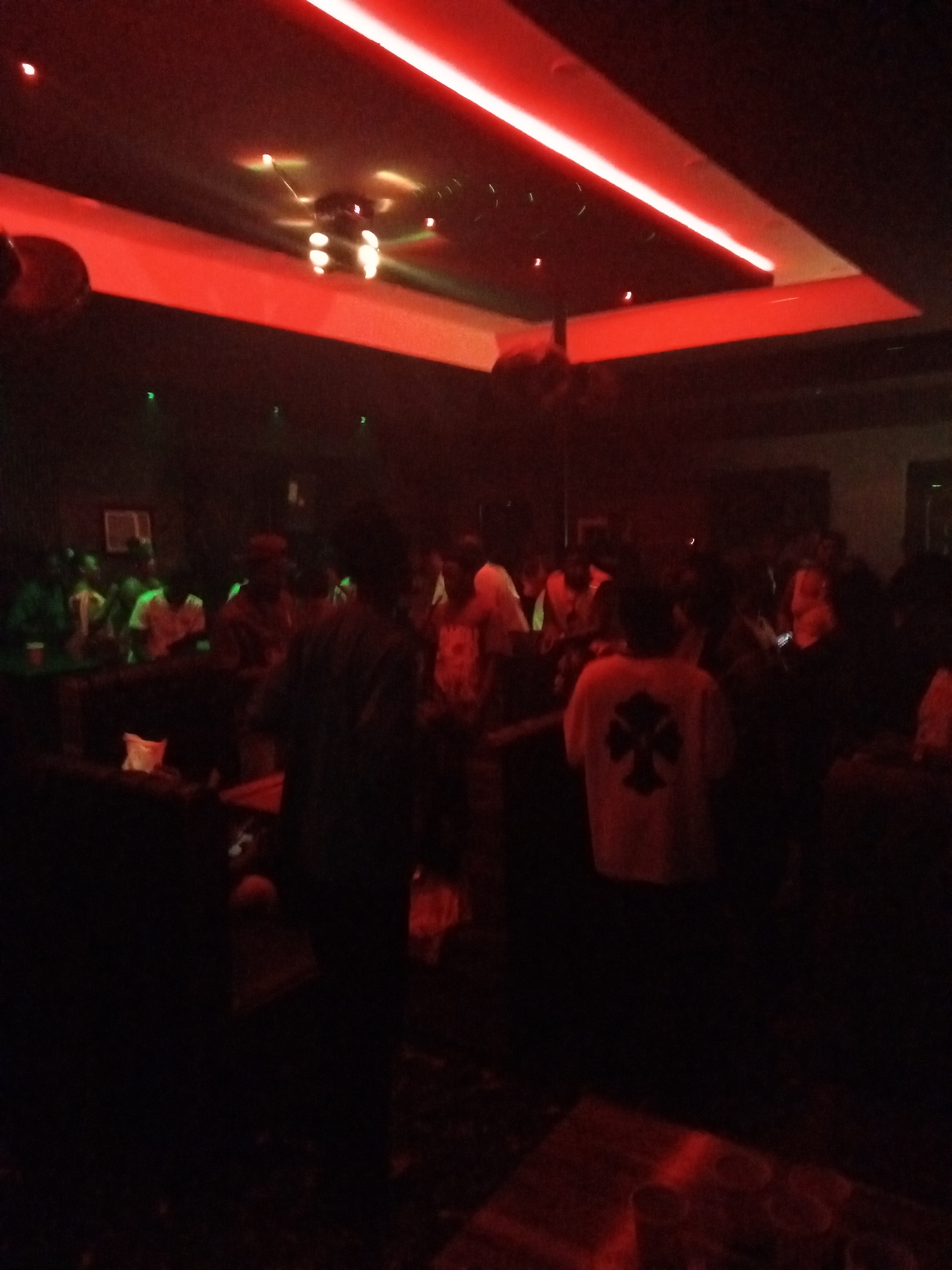 The image shows a dimly lit nightclub with numerous people inside. The location is Unknown, Unknown. Red LED lighting fixtures illuminate the ceiling, casting a reddish hue throughout the scene. A cluster of decorative lights are centrally positioned on the ceiling. Individuals are standing and mingling throughout the space, some near a bar area visible in the background. A person in a white t-shirt with a distinct cross symbol on the back is standing closer to the foreground. Tables and various objects, including what appear to be glasses, are scattered in the foreground. The overall environment is dark and crowded.