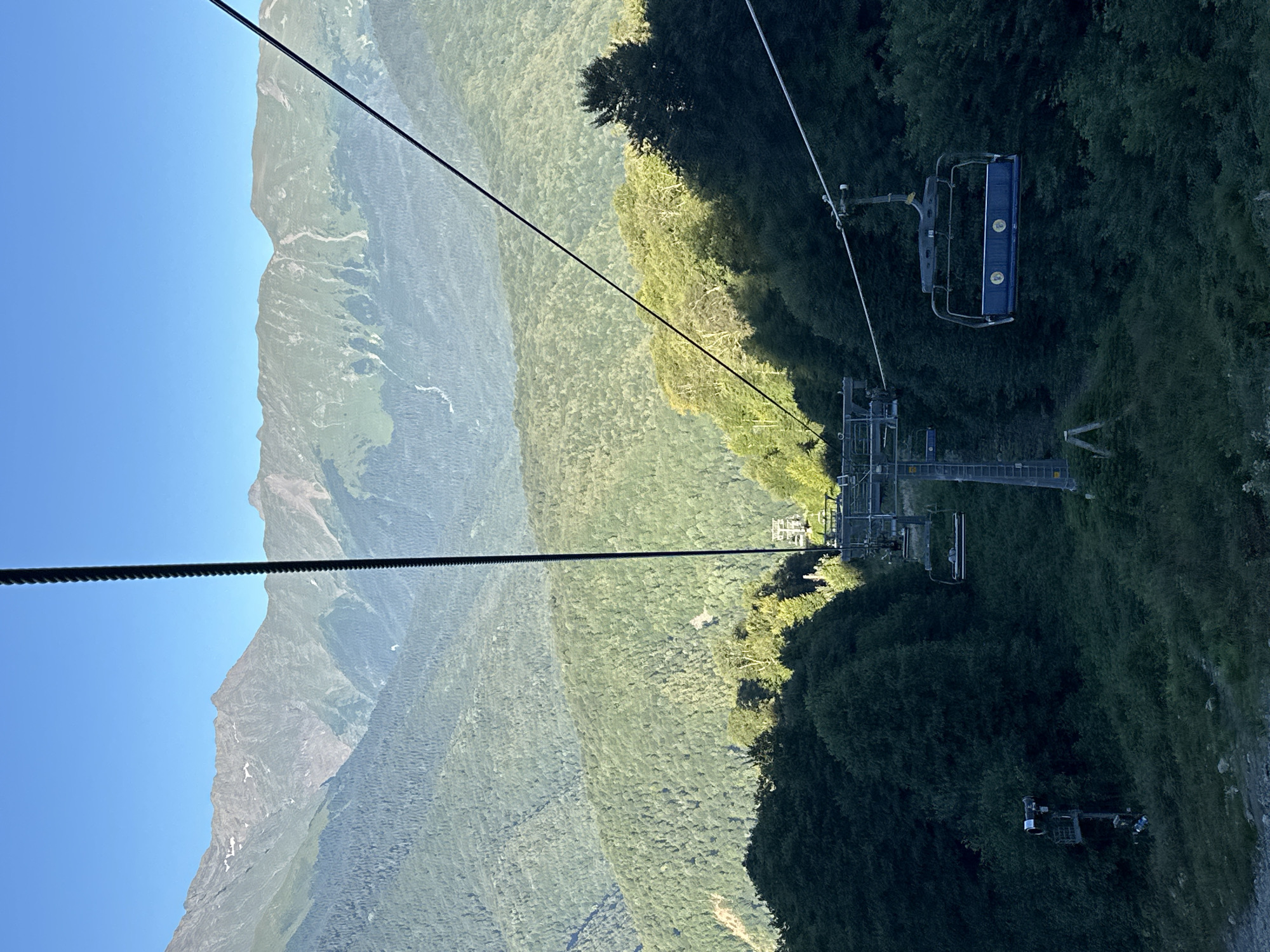 The image depicts a ski lift ascending a forested mountain in Estosadok, Russia. The ski lift consists of blue chairs suspended from a thick cable. The chairs appear to be empty, except for one chair which has two passengers. In the background, layers of green, tree-covered mountains stretch towards a clear blue sky, with rugged, snow-capped peaks visible in the far distance. The sun is shining, casting shadows across the landscape. The ski lift structure itself is visible, with supporting towers and cables stretching upwards. The overall scene suggests a scenic mountain environment, likely during the summer or early autumn, given the lush greenery.