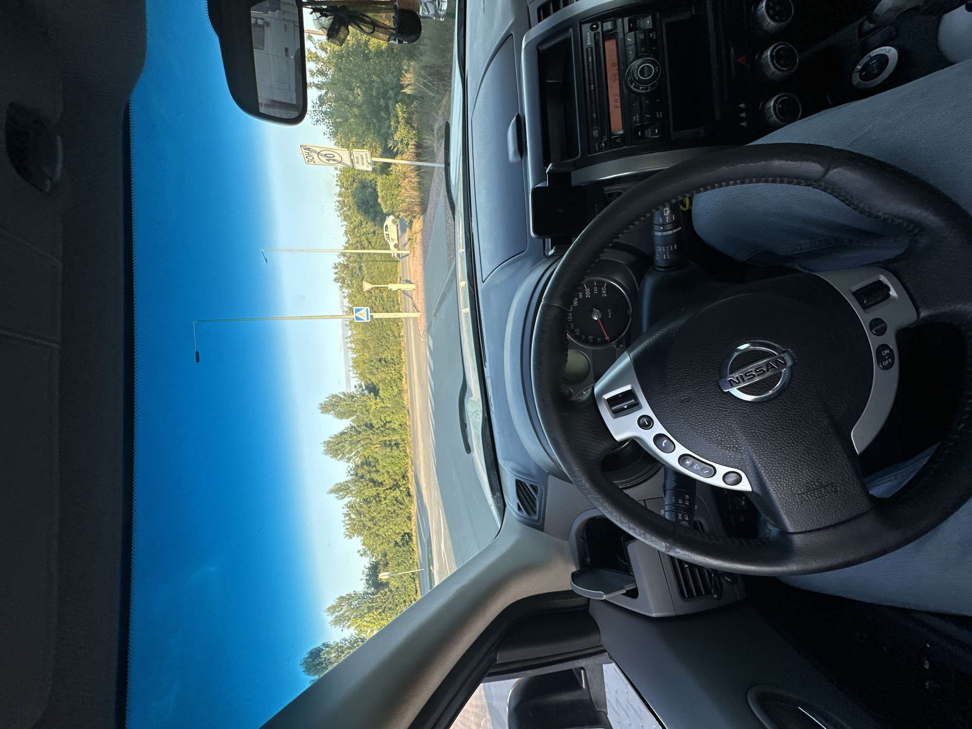 The image is a first-person view from inside a Nissan vehicle, looking forward through the windshield. The interior of the car is visible, including the steering wheel, dashboard with a radio and climate controls, and a part of the driver's leg clad in blue denim jeans. Outside the car, a road lined with trees is visible under a clear blue sky. Road signs are present, including a speed limit sign indicating "10 ZONE" and another with a pedestrian crossing symbol. A white car is parked on the side of the road in the distance. The setting appears to be a suburban or rural area, potentially on the outskirts of Kamianets-Podilskyi, Ukraine, given the presence of road signage that could be consistent with Ukrainian road markings. The lighting suggests it is daytime, likely late afternoon due to the warm, low-angle sunlight.