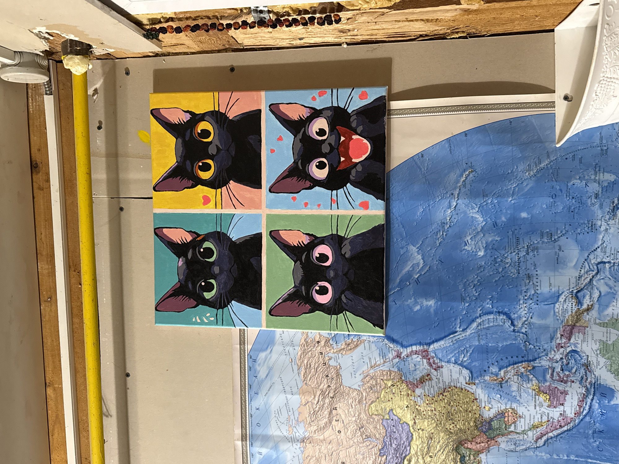 The image displays a painting of four black cats arranged in a four-panel grid. Each cat has a distinct expression and background color: the top left cat has green eyes and a teal background; the top right cat has yellow eyes and a yellow background with a pink heart; the bottom left cat has pink eyes and a green background; and the bottom right cat has a wide-open mouth with a red tongue and a blue background with red hearts. The painting is mounted on a wall. To the left of the painting, a portion of a world map is visible, showing parts of Asia and the Pacific Ocean. The wall itself appears to be in a state of disrepair, with visible screws, a yellow pipe running horizontally, and a section of exposed wood with insulation on the right side. A white container is present in the lower right corner of the image. The lighting suggests an indoor environment, possibly a workshop or storage area, given the unfinished appearance of the wall and the presence of pipes. The location of Sovetskiy, Russia, is not discernible from the visual elements present in the image.