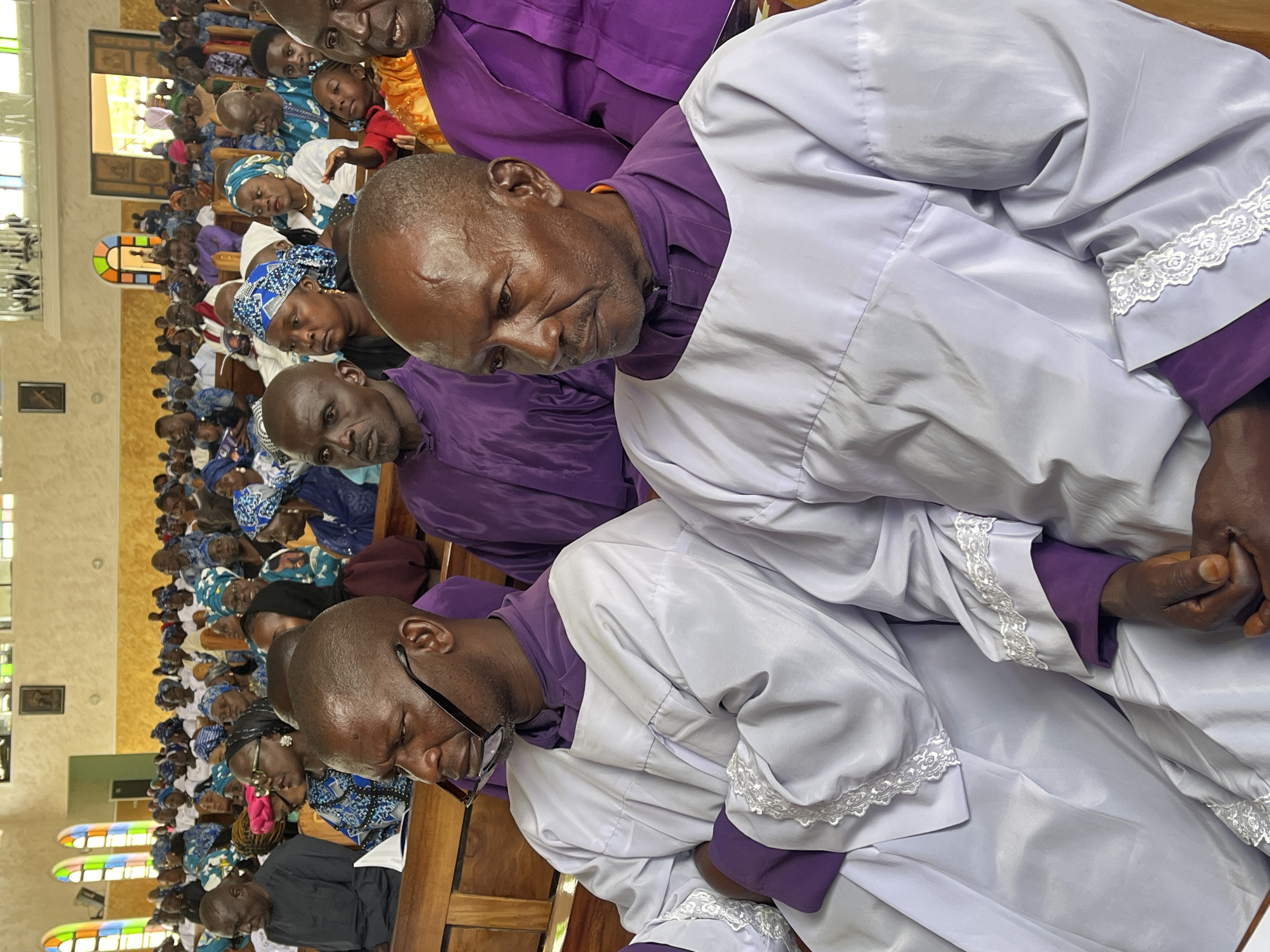 This image depicts an indoor gathering of people in what appears to be a church in Vom, Nigeria. In the foreground, two men are seated, both wearing white robes over purple garments. The man on the left has glasses perched on his nose and is looking to his left. The man on the right is looking straight ahead and has a stern expression. Behind them, a crowd of people, predominantly women, are seated in rows of pews. Many of the women are wearing colorful headscarves. The environment appears to be a church with wooden pews and arched windows with stained glass in the background. The lighting suggests it is daytime.