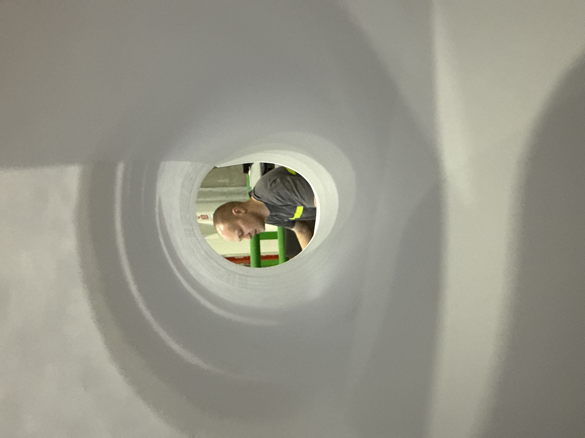 The image provides a unique perspective, looking out from the interior of a large, light-colored tubular structure, possibly a pipe or cylindrical component. The smooth, somewhat reflective surface of the tube dominates the foreground, with subtle concentric rings visible towards the opening.
Through the circular aperture, a person is visible, bent over and looking downwards. The individual, who appears to be a man with a bald head, is engaged in an activity that suggests inspection, work, or maintenance within an industrial or manufacturing setting. He is wearing a dark gray or black short-sleeved shirt with a prominent bright yellow or lime-green stripe along the side or back.
In the background, surrounding the person, parts of green-colored metal structures are discernible, possibly scaffolding, a ladder, or machinery, hinting at a workplace environment. The area beyond the tube appears well-lit by artificial light. To the left of the person's head, a small, indistinct object with a red rectangular shape is visible, which could be part of a sign or electrical component, but no text is legible. This scene takes place in Lupanovo, Russia.