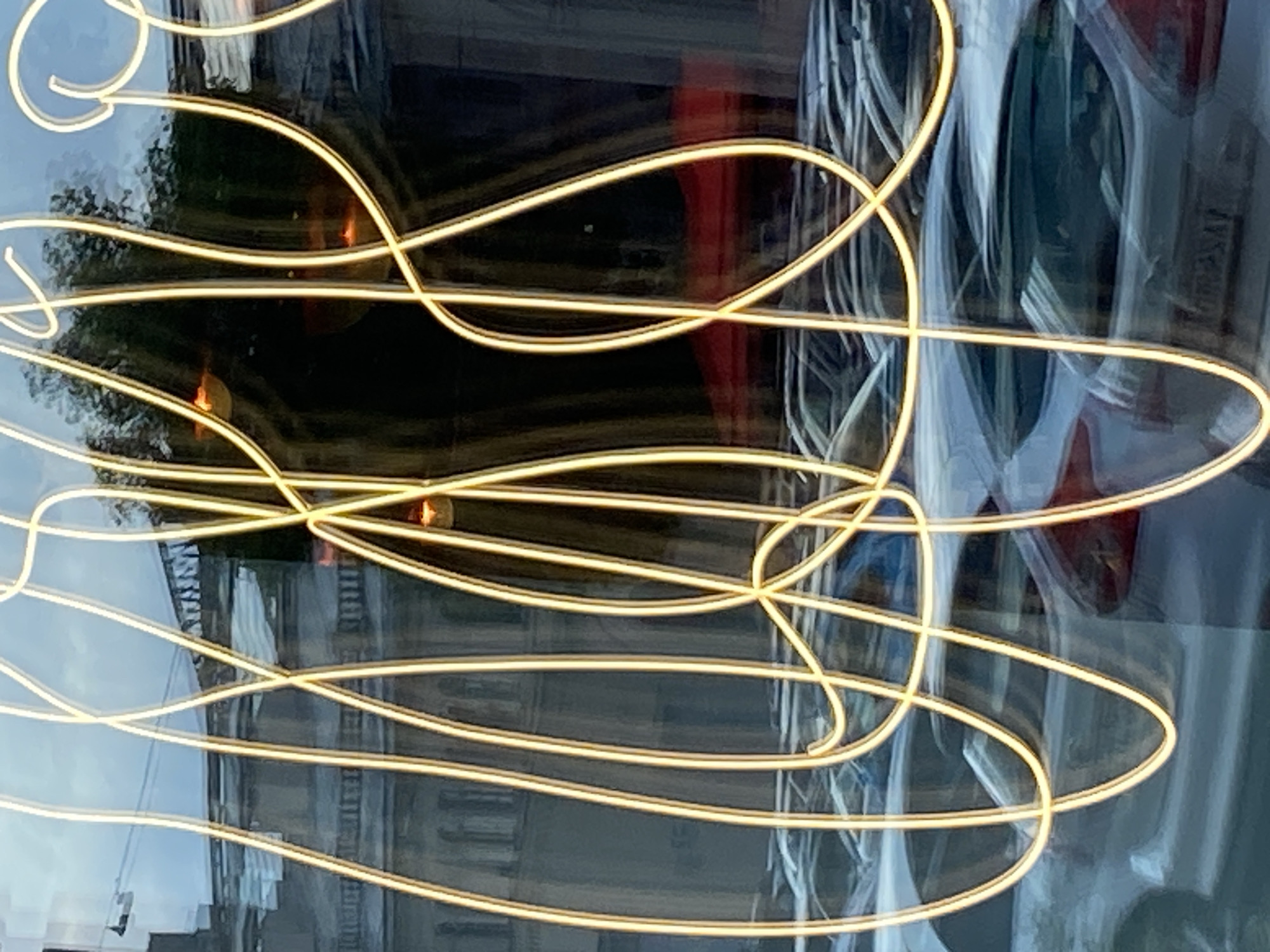 The image captures a reflection on a glass surface, likely a shop window, in Saint Petersburg, Russia. The foreground is dominated by a large, abstract neon light installation in a golden-yellow hue, forming a chaotic, interwoven pattern of lines and curves. Behind this neon, the reflection shows a street scene. Multiple cars are parked in rows, appearing as blurred shapes and outlines due to the reflection and possibly motion. The architecture in the reflection suggests a building with classical detailing, possibly historical, consistent with Saint Petersburg's urban landscape. The sky in the reflection is a pale blue, indicating daytime. There are no visible people.