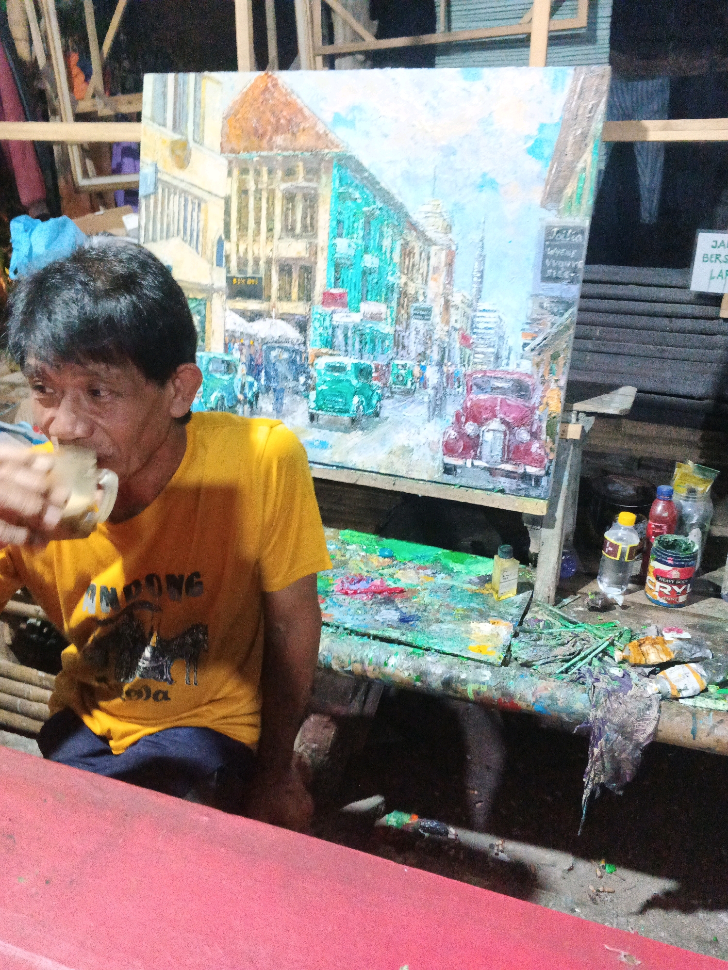 A middle-aged East Asian man in a yellow t-shirt drinks from a clear mug. He is seated to the left of a large, unfinished painting of a street scene with buildings and vintage cars. The painting is on an easel. The man's t-shirt has text and an image printed on it. In front of the painting is a table covered in paint splatters and art supplies, including bottles of paint and brushes. The background shows a rustic wooden structure. The scene appears to be an outdoor art studio or workspace. The presence of the painting, art supplies, and the man's casual attire suggest he is an artist at work. The location is identified as Ungaran, Indonesia.