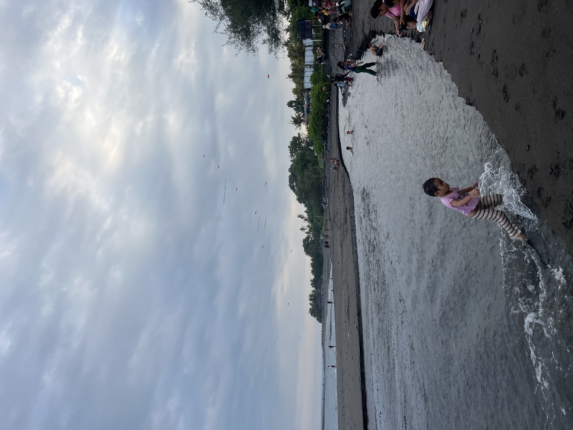 The image depicts a wide, dark sand beach in Blahbatuh, Indonesia, with a river flowing into the ocean. The sky is overcast with scattered clouds. Several people are present on the beach and in the shallow water. In the foreground, a young child wearing a pink shirt and striped pants is playing in the water, splashing with their feet. Further back, other children and adults are visible wading in the river and sitting on the sand. In the distance, there is a line of trees and buildings along the coast. Multiple red kites are flying in the sky. The scene appears to be a recreational gathering on a cloudy day.