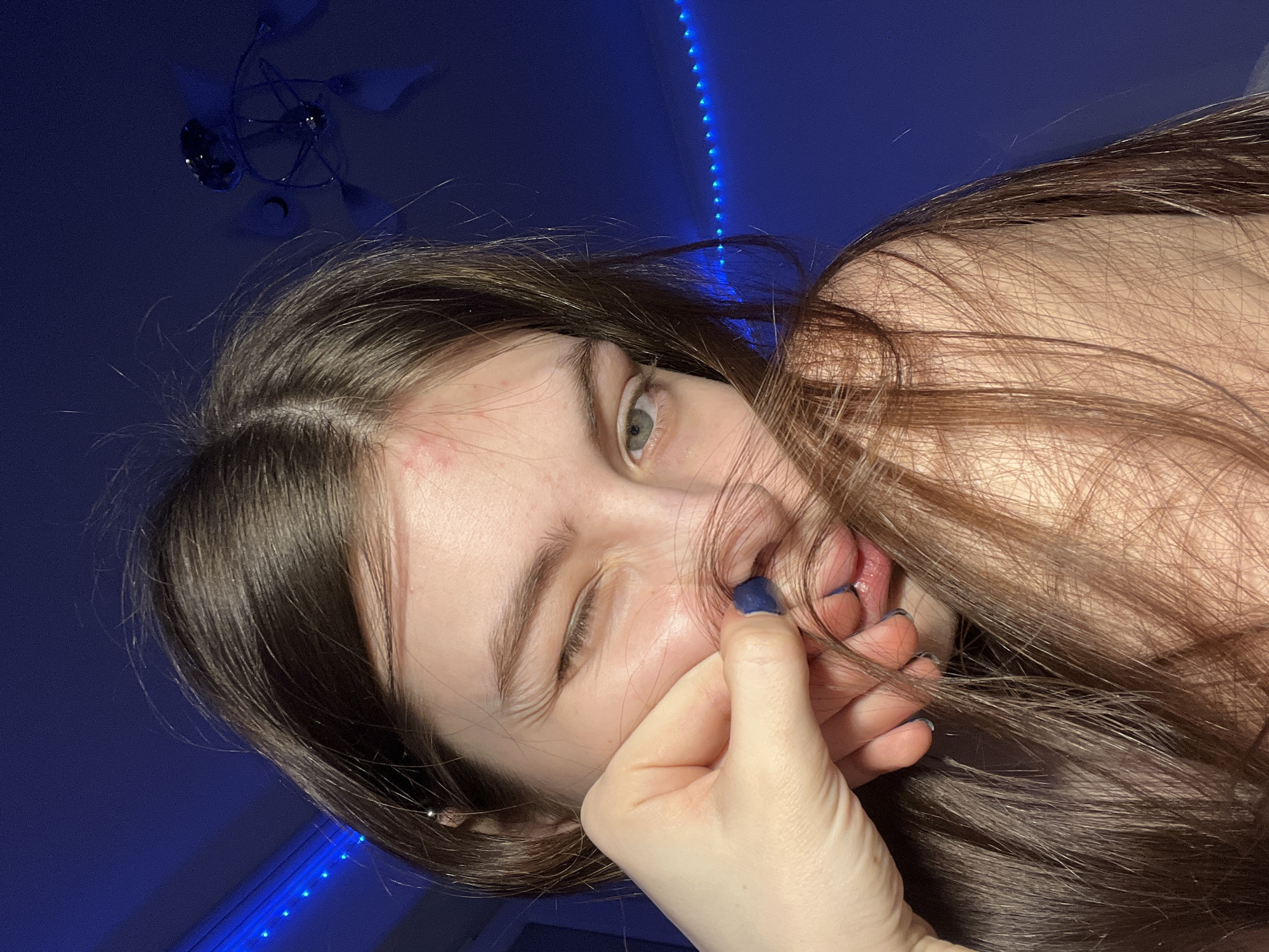 A young woman is shown in a close-up, selfie-style photograph taken indoors under blue lighting. She is winking her left eye and has brown hair partially covering her face. Her hand is near her mouth, with some strands of her hair pulled between her fingers and lips. There is a blemish visible on her forehead. In the background, a ceiling fan and blue LED strip lighting are visible. The location is not identifiable as Saint Petersburg, Russia, from the provided image.