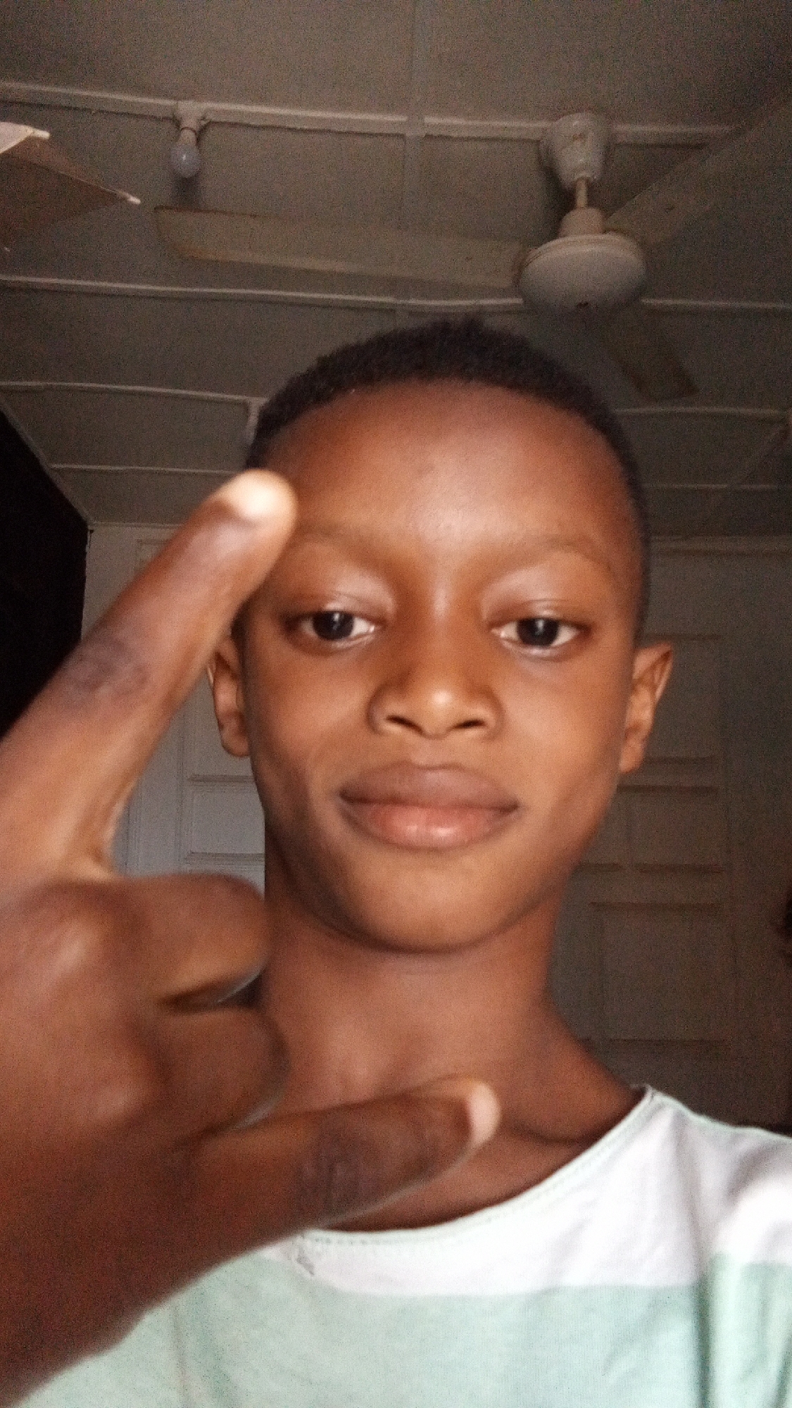 A young individual, appearing male, is captured in a close-up, eye-level indoor shot. They have short, dark hair and dark skin, looking directly at the camera with a neutral to slightly smiling expression. Their left hand is raised in the foreground, forming a gesture with the index finger pointing upwards and the pinky finger also extended, while the middle and ring fingers are bent down. A faint discoloration or mark is visible on their index finger.
The person is wearing a t-shirt with a white or light-colored upper section and a light green or teal lower section. The background features an indoor setting with a low ceiling made of light-colored panels or planks. A white ceiling fan is visible in the upper right, and a bare light bulb hangs from the ceiling in the upper left. A wall or door frame is faintly seen behind the person's head. The lighting is somewhat dim, suggesting an indoor environment possibly relying on artificial light. The image appears to be a selfie, capturing a casual moment indoors in what is contextually identified as Sabon Tasha, Nigeria. There is no visible text in the image.