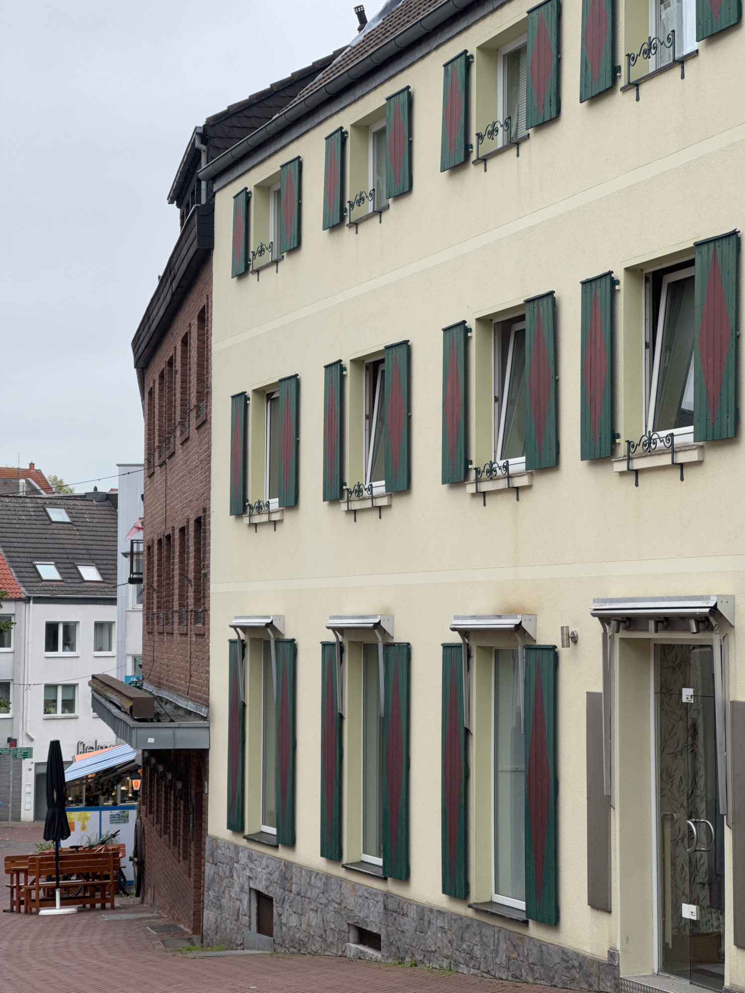 The image shows a view of the facade of a building in Cleves, Germany. The building has a light yellow stucco exterior with several windows. Each window is fitted with dark green shutters that have a diamond pattern in a dark red color. The shutters are in various states of being open or closed. The building has at least three stories visible. On the ground floor, there is a glass door with stainless steel handles. The building is situated on a brick-paved street. To the left of the main building, a brick building is partially visible. There are benches and a black umbrella set up on the sidewalk in front of the brick building, suggesting an outdoor seating area. In the background, other buildings and a cloudy sky are visible.