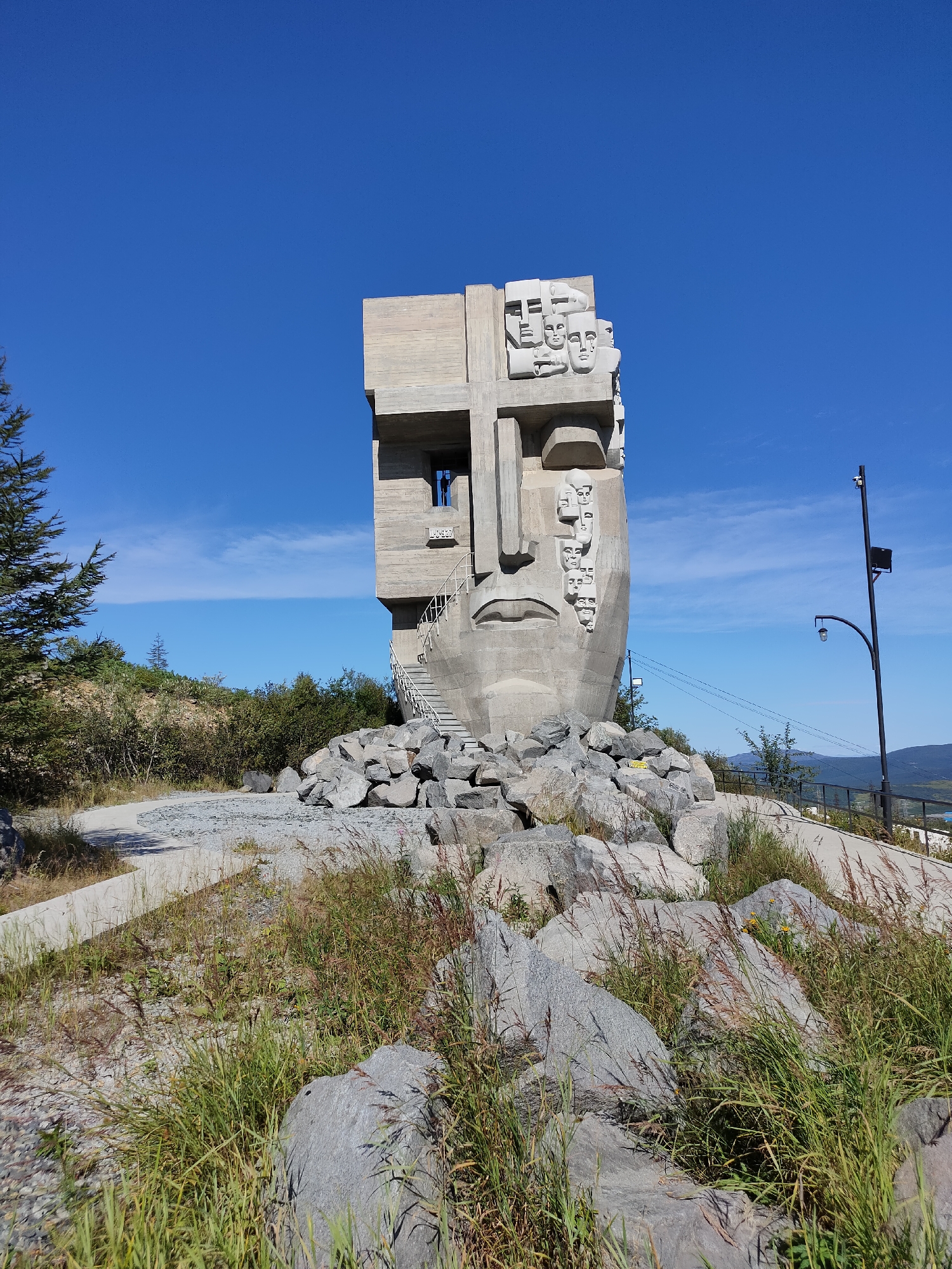 This is a full-shot photograph of the "Mask of Sorrow" monument in Magadan, Russia. The monument, a large concrete sculpture of a human face with a sorrowful expression, is situated on a hilltop overlooking a landscape of hills and sparse vegetation under a clear blue sky with a few wispy clouds. The sculpture is surrounded by large rocks and patches of dry grass and weeds. A white spiral staircase is visible ascending the side of the monument, leading to an opening resembling a window. In the background, a lamppost with a light fixture stands to the right of the monument, and a black metal railing lines a pathway that curves away from the sculpture. The overall scene depicts an outdoor monument in a somewhat desolate, natural setting.
