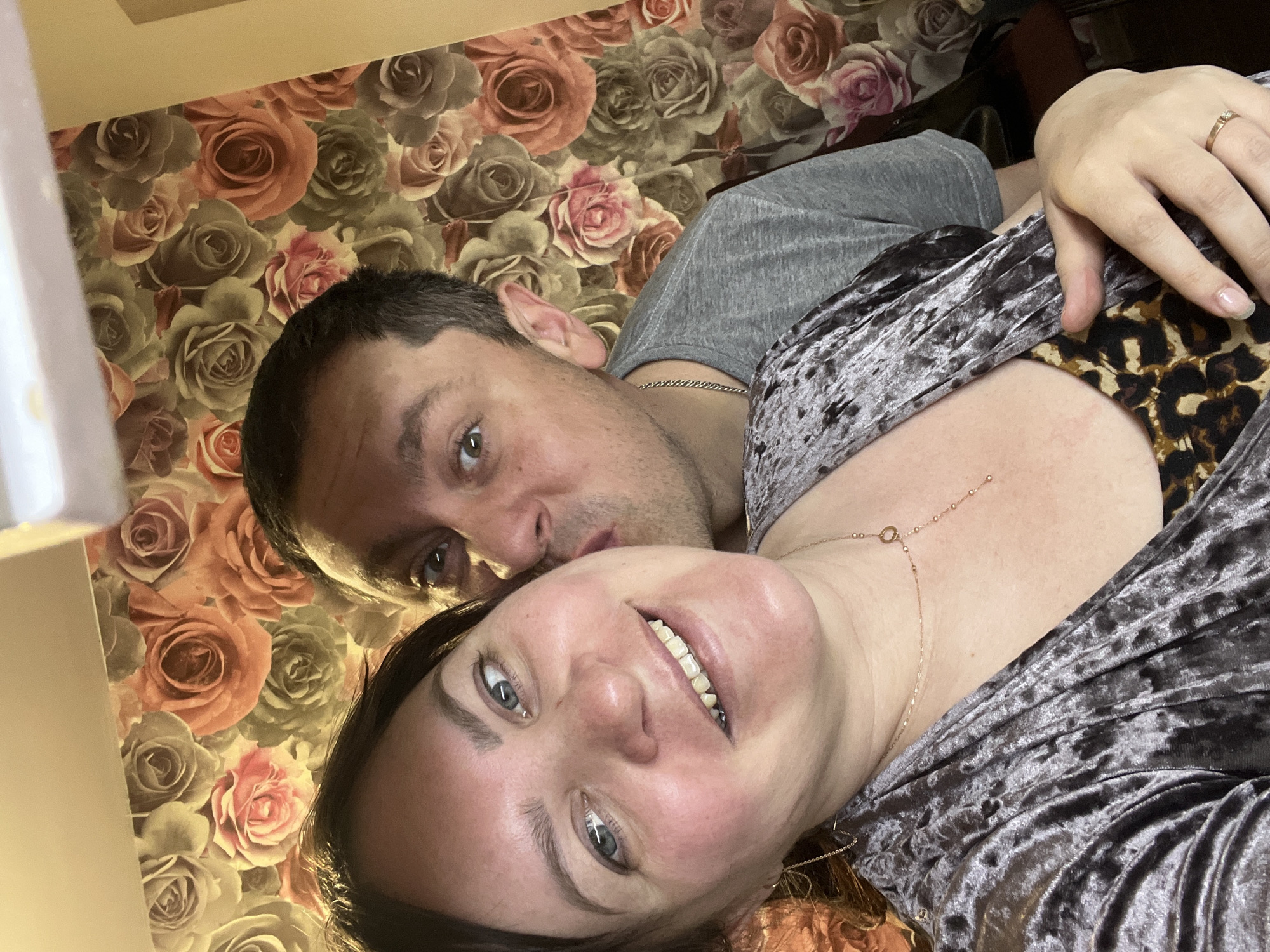 A man and woman are posing for a close-up selfie. The woman is in the foreground, with her face angled towards the camera and a visible smile. She is wearing a grey velvet top with a leopard print portion visible at the neckline and a delicate gold necklace. The man is positioned behind her, wearing a grey t-shirt and a chain, with his face close to hers. The background features a wallpaper with a rose pattern in shades of brown, peach, and pink. The scene is lit from the left. The location is unknown.