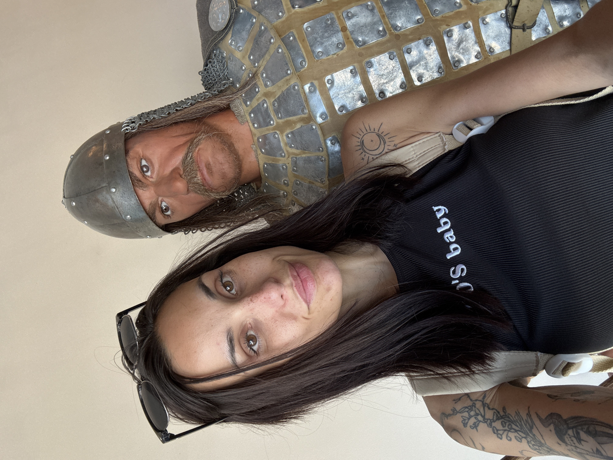 A dark-haired woman poses for a selfie with a mannequin dressed in medieval armor. The mannequin has long brown hair, a helmet, and a beard. The woman is wearing a black shirt and sunglasses. The scene is likely indoors, with a light beige or white background. A tattoo of a sun is visible on the woman's arm. This photograph was taken in the city of Veliko Tarnovo, Bulgaria.