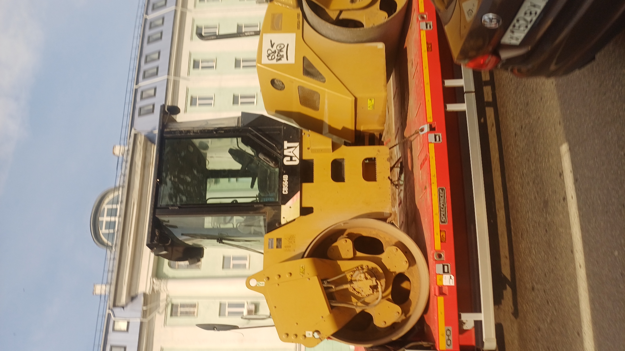 In Izhevsk, Russia, a yellow Caterpillar CS664D tandem vibratory roller is transported on a red flatbed trailer. The roller features a prominent cab with tinted windows and visible interior controls. The front drum of the roller is large and robust, with an intricate hub assembly. The trailer itself displays the branding "SPEC PRICE P". In the background, a multi-story building with light-colored facades and distinctive arched windows is visible. The sky above is clear with a light blue hue. The scene appears to be a street in the city, with a portion of a dark-colored passenger car visible in the foreground on the right, its license plate partially obscured. The overall image captures a moment of transportation of heavy construction equipment within an urban environment.