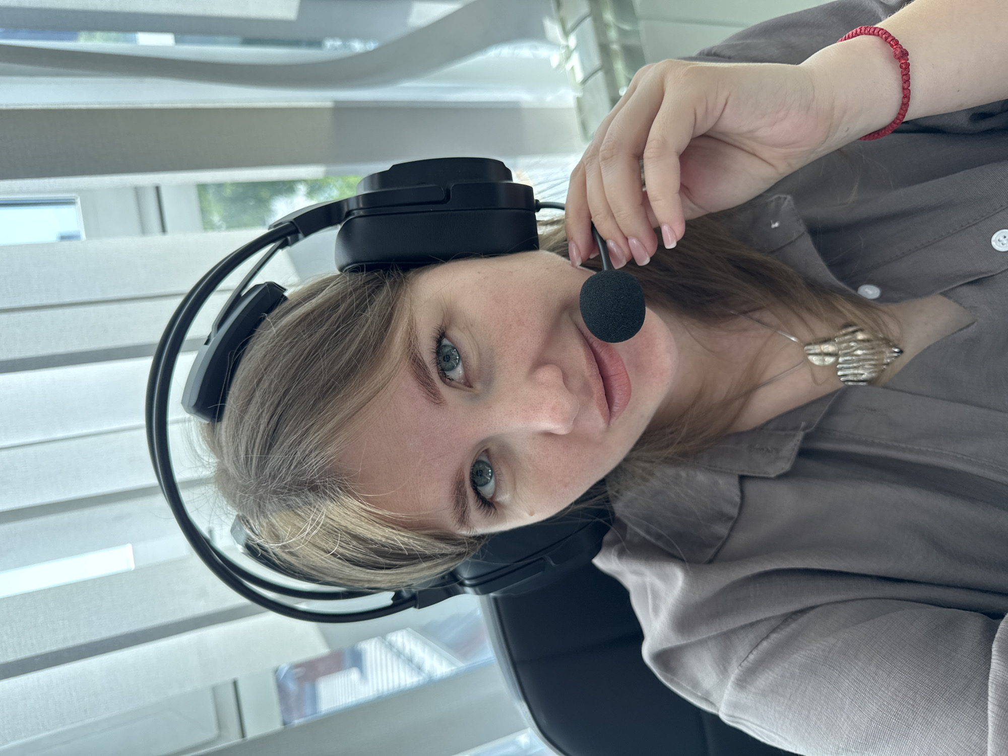 A fair-skinned woman with light blonde hair and blue eyes is wearing a black headset with a microphone and a gray button-down shirt. A silver necklace is visible. Her fingernails are manicured with a light pink polish. She is wearing a red string bracelet on her right wrist. The background features a window with white blinds, suggesting an indoor setting. The image was likely taken indoors in the city of Perm, Russia, based on the user request.