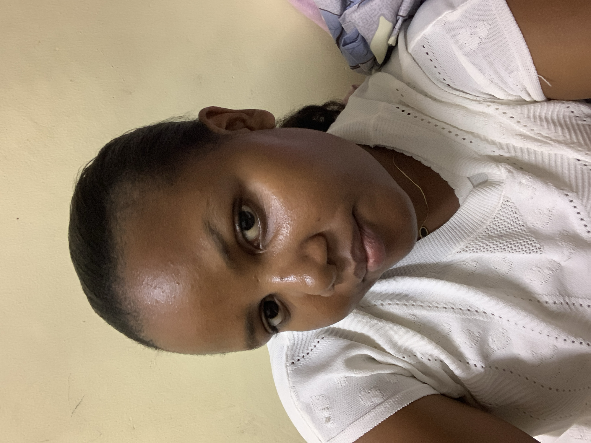 A dark-skinned woman with short black hair is positioned in a vertical orientation against a pale yellow wall. She is wearing a white knit top. A gold chain is visible around her neck. A partially visible sheet of blue, white, and purple fabric is located in the upper right corner of the image. The image does not provide enough information to determine a specific location in Dar es Salaam, Tanzania.