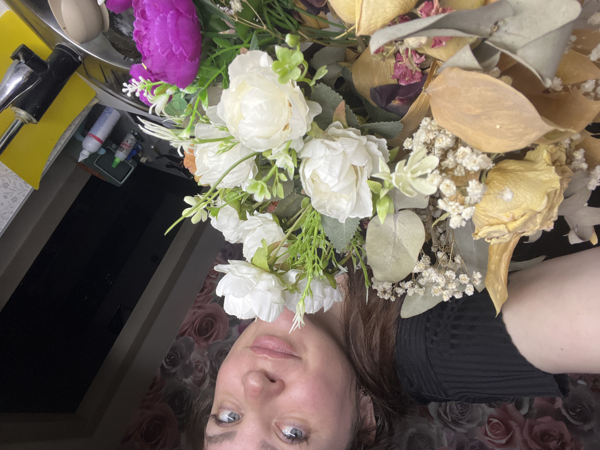 An indoor, close-up, selfie-style image captures a woman and a floral arrangement within a residential setting in the city of Noviy, Russia.
The upper right portion of the frame features a female individual from the chest up, identified by her light skin tone, blue eyes, and medium-brown hair. She wears a black, horizontally ribbed top and displays a slight smile while looking directly at the camera. A small mole is visible on her left shoulder, positioned on the left side of the image.
The left and central-lower areas of the image are dominated by a mixed floral display. This arrangement includes large, dried, light-yellow flowers; numerous small, white dried florets resembling baby's breath; flat, tan-colored dried leaves or pods; and clusters of dried pink petals. Interspersed are several prominent artificial white peonies with green foliage, and a large, artificial purple peony is visible in the lower right quadrant. Grey-green dried eucalyptus-like leaves are also part of the arrangement.
The background to the upper left features wallpaper with a repeating pattern of muted pink and grey roses. To the right, a domestic sink area is visible. This includes a metallic sink basin, a black faucet, and a bright yellow mat or sponge placed on the right side of the sink. Behind the sink, on a dark countertop, are two bottles: a larger white bottle with a red component and a smaller bottle with a green cap. A light-colored spoon rests within the sink basin. Above the sink, dark, angled architectural elements are present.