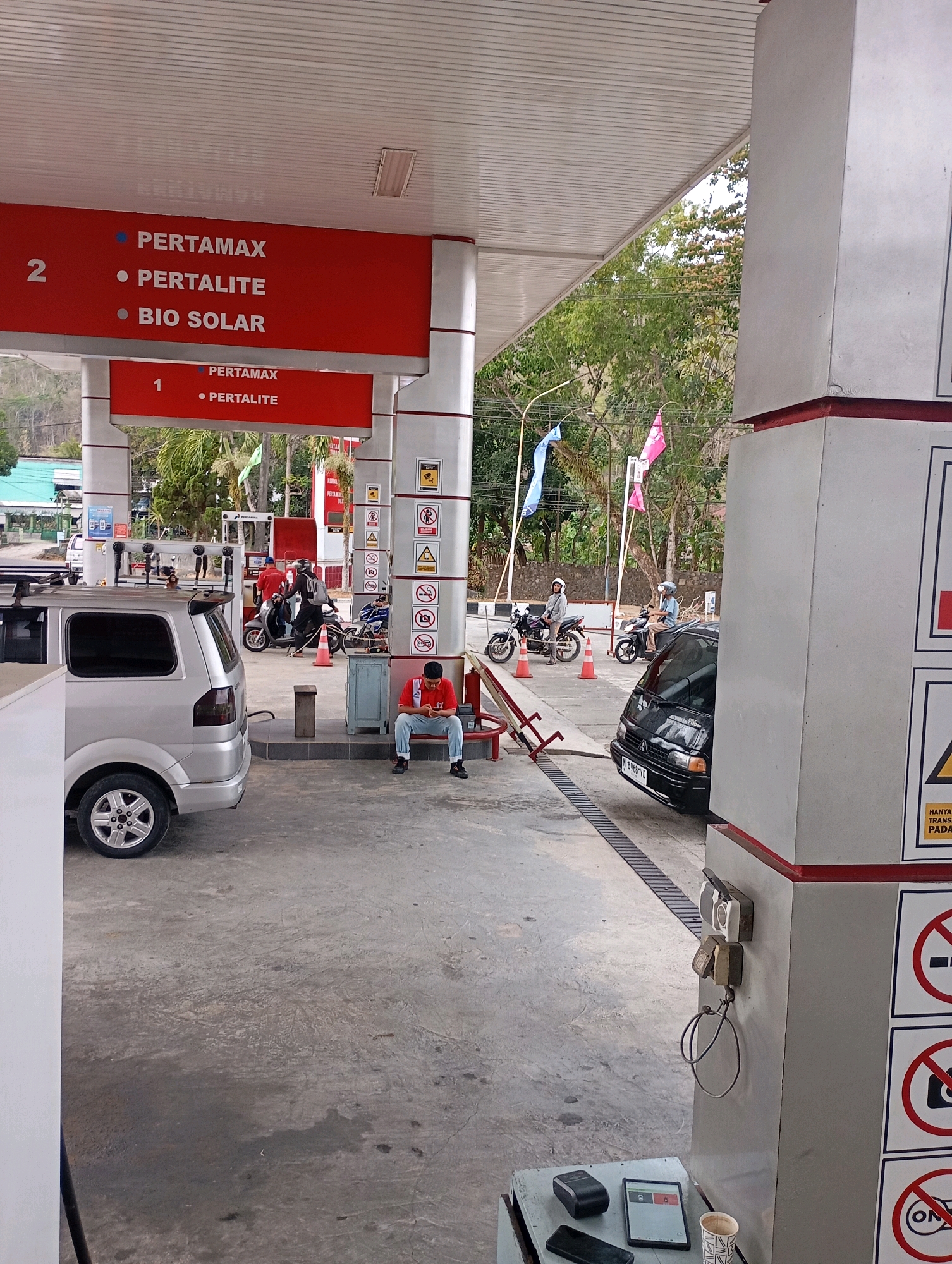 A man in a red shirt and blue jeans sits on a low concrete step at a gas station in Menadi, Indonesia, looking at his phone. To his left, a silver minivan is parked. In front of him, a black car is parked at a gas pump. Several motorcycles are also present, with people on them. The gas station has a red and white awning and signs listing different fuel types like "PERTAMAX," "PERTALITE," and "BIO SOLAR." The ground is made of concrete, and trees and foliage are visible in the background. The sky is bright, suggesting a daytime scene. There are also safety signs and a CCTV camera visible on one of the station's pillars.