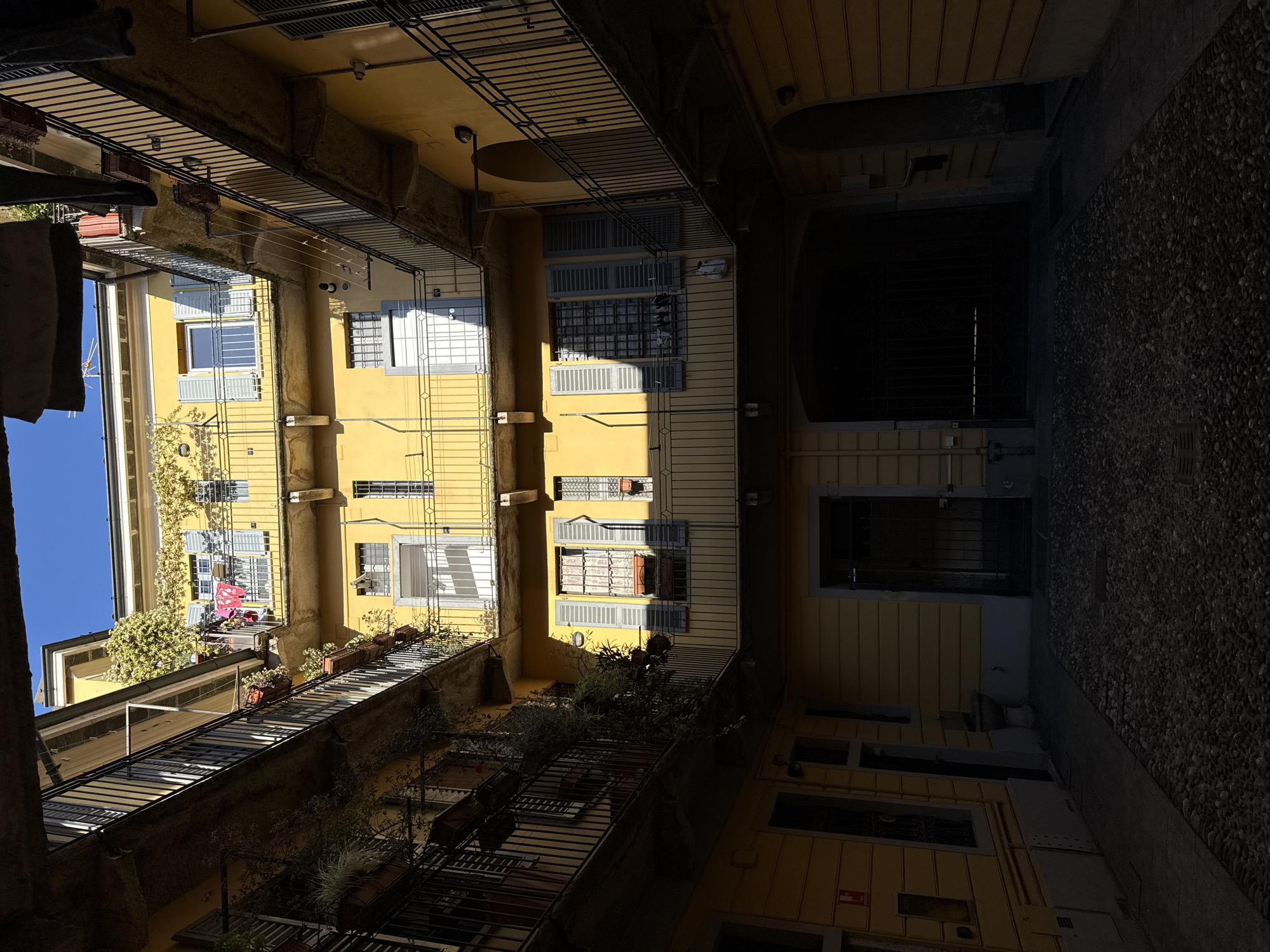 This is a low-angle shot looking up within an urban courtyard or narrow street, likely in Milan, Italy, suggested by architectural styles. The focal point is the buildings lining the space, particularly one painted yellow with multiple windows, some with closed shutters. Architectural details include iron balconies and window bars. Hanging clothes can be seen in one area. The right side of the frame shows a dark, shadowed passage or entrance, with what appears to be a textured wall. The lighting is uneven, with some areas brightly lit and others in deep shadow, suggesting a time of day with strong sunlight. There are no visible people or animals. The image primarily captures the architecture and spatial arrangement.