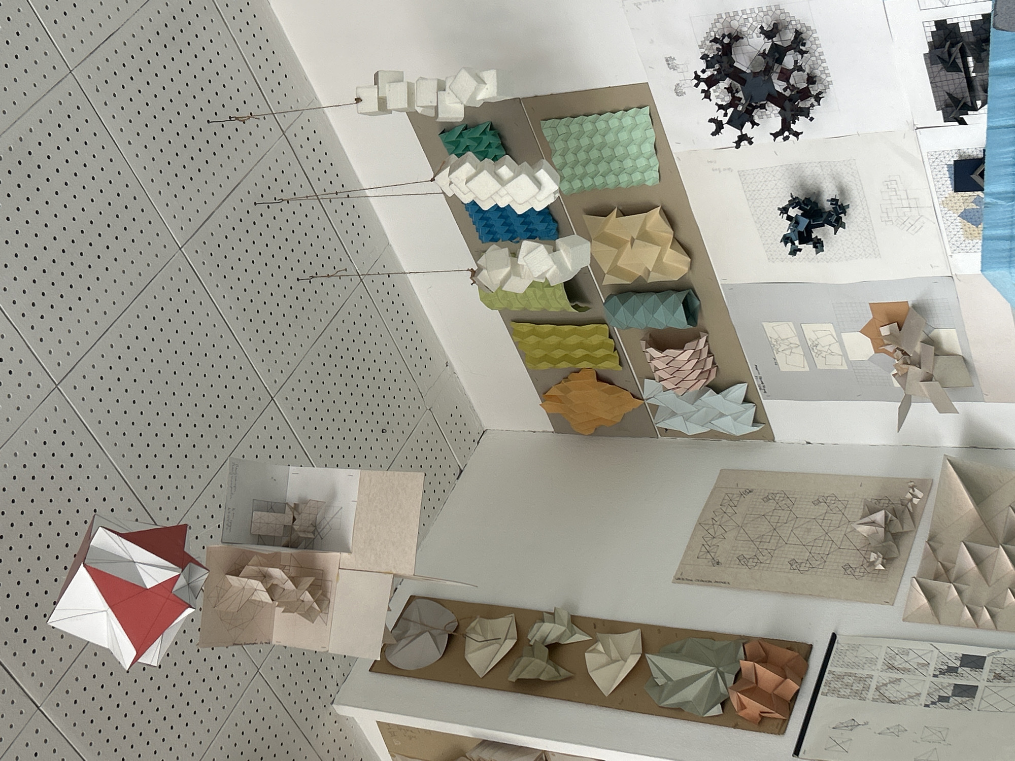 This media file captures an overhead, angled view of a detailed display showcasing various architectural or design models and studies, likely in a studio or exhibition space in Sofia, Bulgaria. No people are visible.
The scene features a diverse collection of geometric paper models, sketches, and design iterations. On the left, a light grey perforated wall or ceiling serves as a backdrop, from which several series of white, cuboid paper models are suspended by thin wires. Below these, additional folded paper models in teal, blue, light green, and olive green are arranged on a brown board, exhibiting different tessellation and pleating techniques.
To the right, numerous sketches and technical drawings on paper are spread across a white surface. Among these are two prominent, dark, intricate fractal-like 3D models, suggesting complex geometric studies. Further down, a large sheet displays a complex hexagonal grid pattern with small folded paper forms placed upon it, alongside other smaller diagrams and sketches.
At the bottom-center and right, a white shelf or display surface holds more folded paper models in muted tones like grey, white, green, and terracotta, some arranged on a brown backing board. These models explore various geometric shapes and folding methods. On the bottom-left, a striking red and white multi-faceted geometric paper model rests directly on the perforated surface, surrounded by smaller, beige-colored folded paper studies.
The display emphasizes modularity, intricate folding techniques, and geometric patterns, suggesting a focus on parametric design, origami, or architectural skin studies. The lighting appears even, indicating an indoor setting. Any visible text on the drawings is too small and indistinct to be legible.