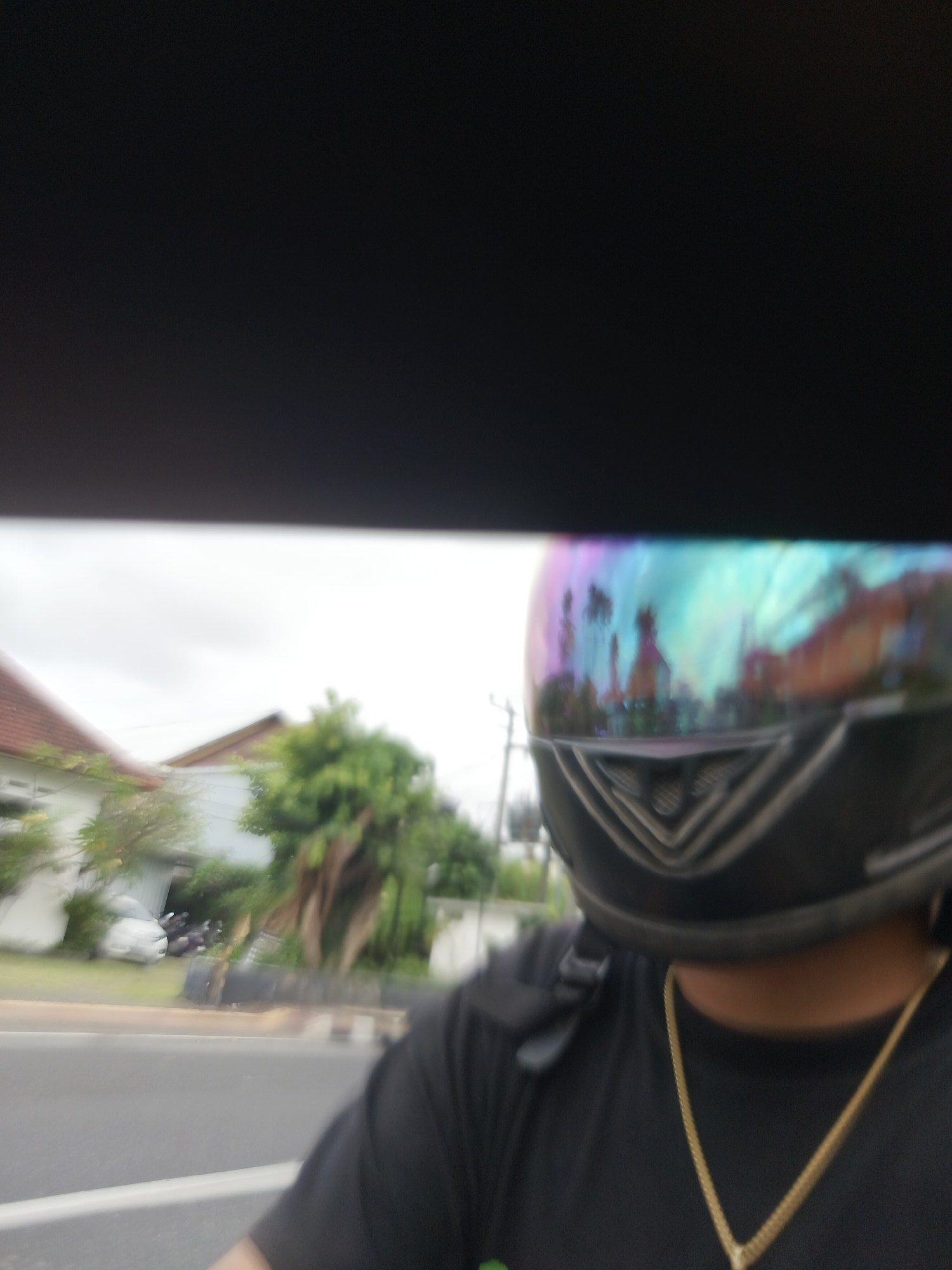 The image depicts a first-person perspective of someone wearing a black t-shirt and a helmet with a rainbow-colored reflective visor. A gold chain is visible around the person's neck. In the background, a street with a white line marking is visible, along with buildings and trees, suggesting an outdoor urban or suburban environment. The sky is overcast. The reflection on the visor shows a blurry depiction of buildings and palm trees, hinting at a tropical location. The presence of motorcycles parked in front of a building in the background further suggests a populated area. The overall scene is consistent with a street view in Denpasar, Indonesia, given the architectural style of the buildings and the vegetation.