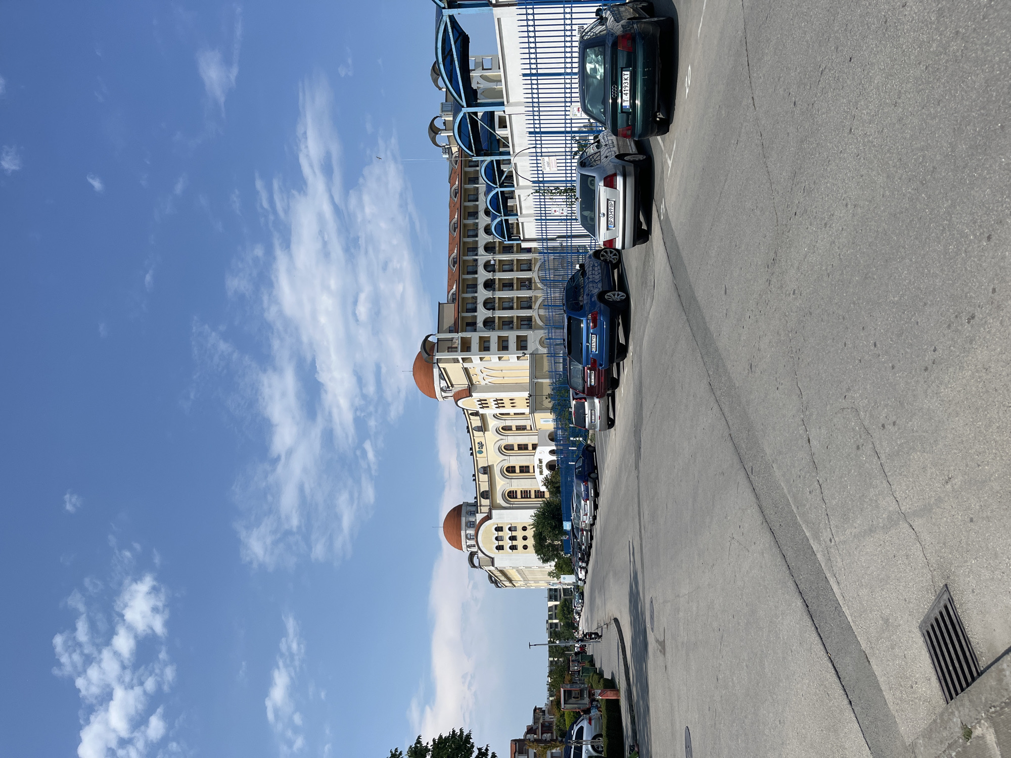 This image captures a street scene in Obzor, Bulgaria, with a prominent building featuring red domes in the background. The building is large and multi-storied, with a pale yellow facade, arched windows, and balconies. A blue metal fence runs along the front of the building, enclosing a parking area. Several cars are parked in a row on the asphalt street. The sky is blue with scattered white clouds. To the left of the street, there are trees, a sidewalk, and some smaller buildings. The overall impression is that of a sunny day in a coastal town.