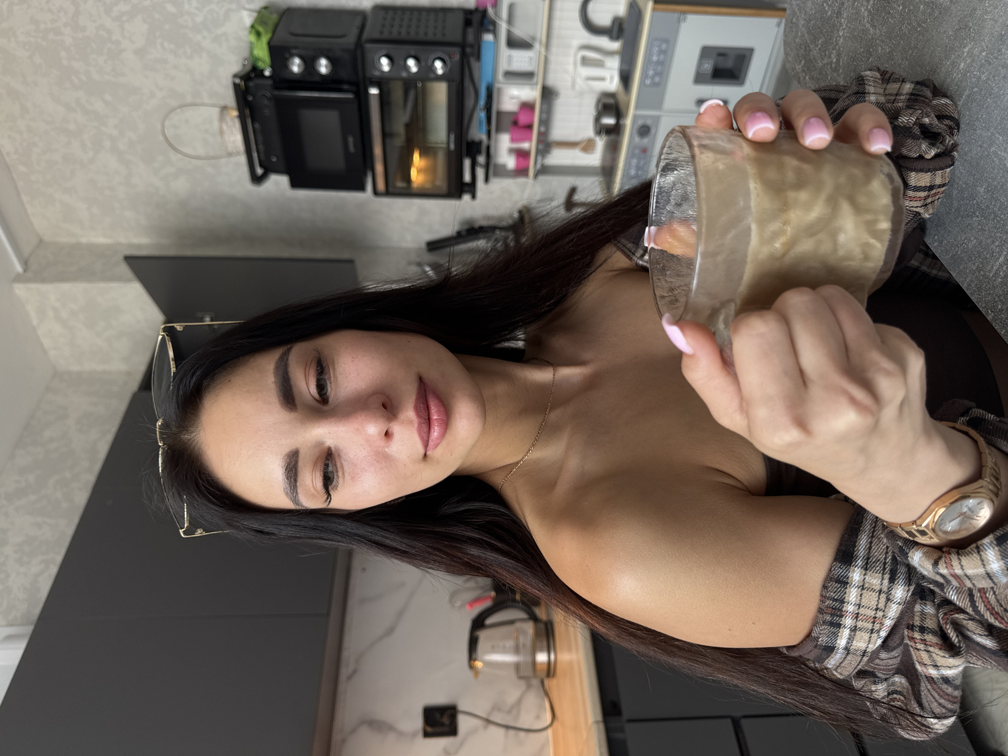 A woman with long, dark hair is holding a glass of a light brown liquid, possibly a beverage, in front of her. She is wearing a plaid shirt and a gold watch. There are glasses on her head and a gold necklace around her neck. The background includes kitchen appliances, such as an oven and a microwave, and a gray kitchen counter. A kettle is visible on the counter. The location is likely a kitchen in an apartment or house. It is not possible to determine if the location is in the city of Chaikovsky, Russia, based on the image alone.