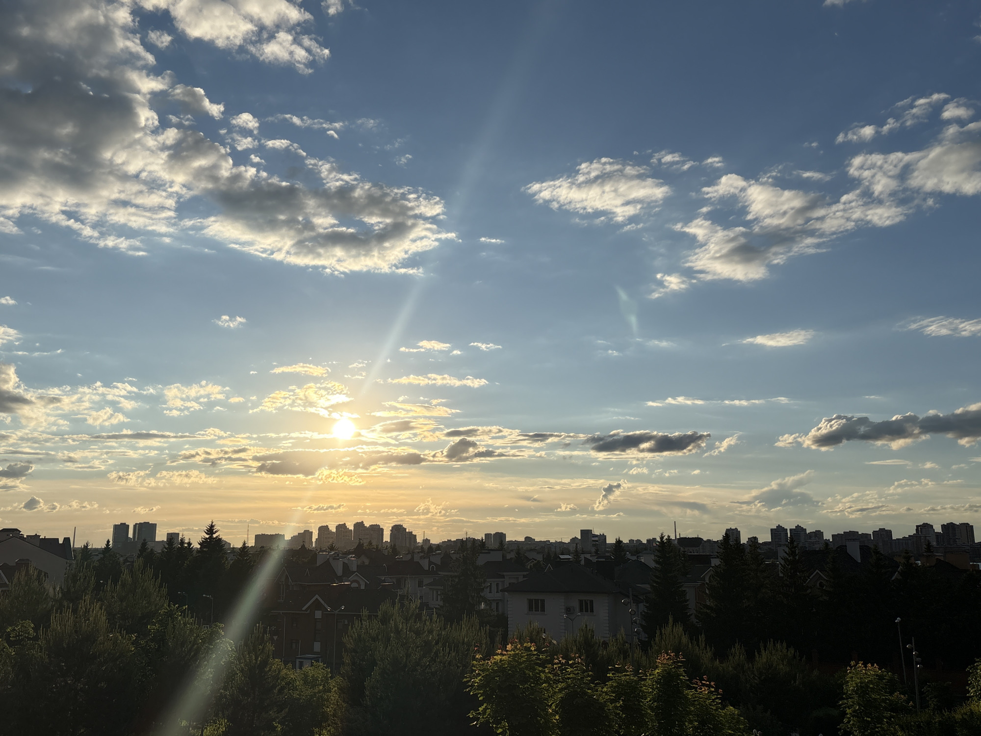This image captures a panoramic view of a landscape at either sunrise or sunset, characterized by a vibrant sky and a mix of natural and urban elements.
The sky dominates the upper portion of the frame, painted in shades of blue and gold. Scattered cumulus clouds are visible, some illuminated brightly by the low sun, creating a warm, golden glow, while others appear darker against the lighter sky. The sun is a prominent feature on the left side of the image, appearing as a bright, circular orb low on the horizon, casting strong rays of light that create noticeable lens flares across the image. These light rays extend diagonally downwards from the sun and also appear as subtle streaks elsewhere in the sky.
Below the sky, the landscape transitions into a lush, green canopy of trees that fill the mid-ground. These trees, mostly coniferous and deciduous, form a dense layer, partially obscuring the residential-style buildings nestled among them. The houses feature pitched roofs and are generally light-colored, suggesting a suburban or residential area.
In the distance, a distinct city skyline is visible, characterized by numerous tall buildings and skyscrapers that appear as dark silhouettes against the brighter sky. This suggests the view is from a higher vantage point, overlooking a metropolitan area with surrounding greenery.
The overall impression is one of tranquility and natural beauty, highlighting the contrast between the warm, golden light of the sun and the cooler blues of the sky, along with the blend of dense foliage and urban structures. There is no visible text in the image.