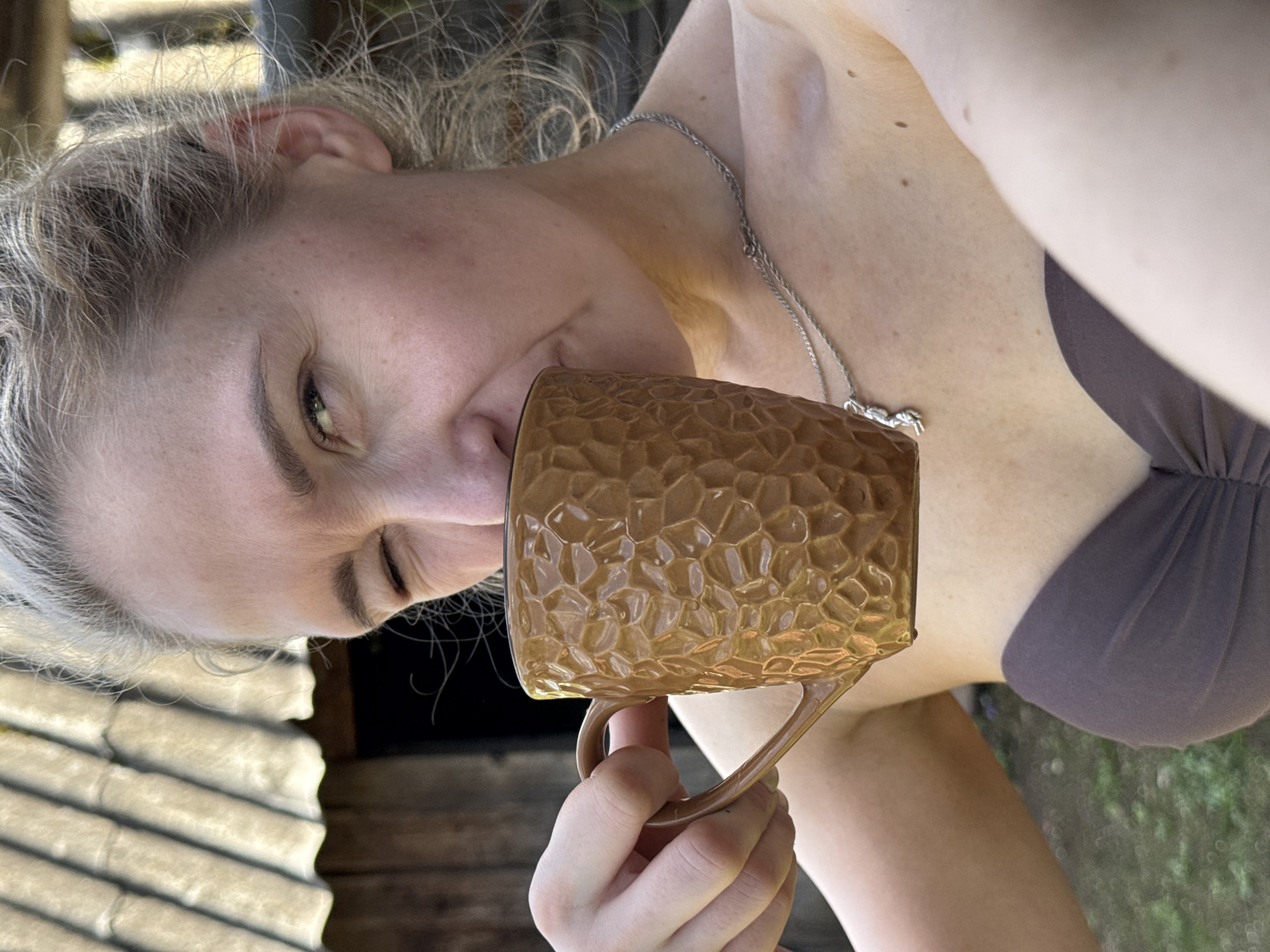 A woman is holding a brown textured mug to her lips. She has blond hair, fair skin, and is wearing a silver necklace and a brown top. The background includes a wooden structure and greenery. The location is not discernible from the image.