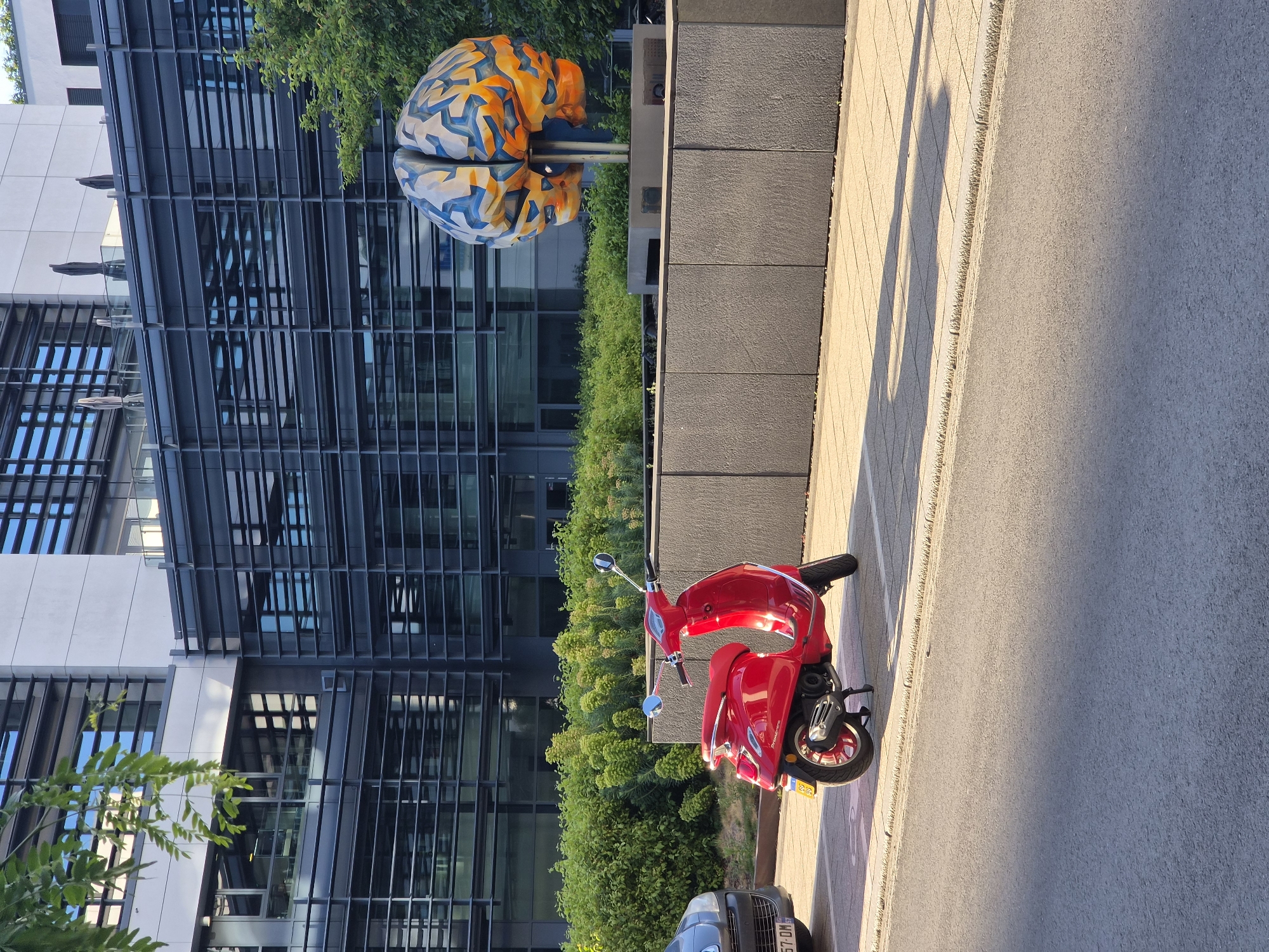 The image depicts an outdoor scene in Sanem, Luxembourg, illuminated by low-angle sunlight, suggesting either early morning or late afternoon. A vibrant red Vespa-style scooter is the prominent subject, parked on a light grey paved surface, facing slightly to the left with its right side visible. The scooter features chrome handlebars, mirrors, and trim, with a small yellow license plate affixed to its rear fender.
To the left of the scooter, the front bumper and grille of a dark grey vehicle, displaying a Luxembourgish license plate partially visible as "57-DM," are present. In the mid-ground, a large, artistic sculpture of a human brain stands on a dark grey concrete pedestal. The sculpture is rendered in a mosaic-like pattern of blue, orange, and light grey geometric shapes.
Behind the scooter and surrounding the brain sculpture, dense green foliage, comprising shrubs and small trees, forms a landscaped barrier. In the background, modern, multi-story buildings with extensive glass facades and dark horizontal louvers are visible. These structures feature light grey exterior paneling. A branch with green leaves partially intrudes into the top-left corner of the frame. The immediate foreground consists of an asphalt road, transitioning to a light-colored concrete sidewalk or paved area. Long, distinct shadows are cast across the ground from the right side of the frame. No individuals are present in the scene.